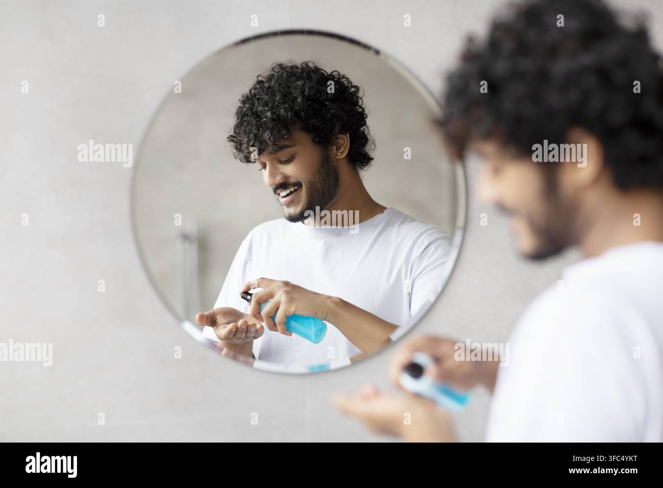 Giovane uomo con capelli ricci che applica un prodotto per la cura davanti allo specchio del bagno, espressione felice Foto Stock