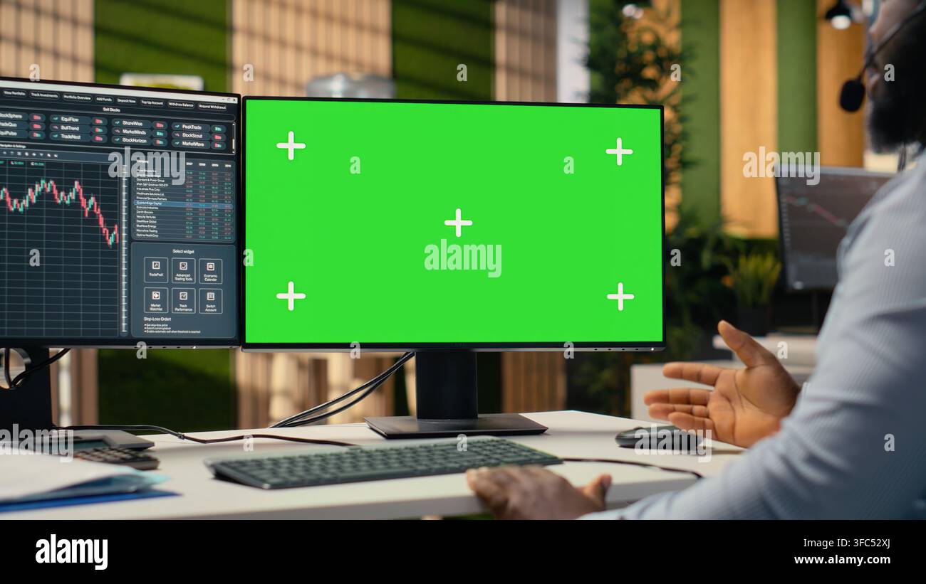Consulente finanziario che utilizza un computer con schermo verde offre consulenza sugli investimenti durante le chiamate con i clienti. Uomo afroamericano che parla in un microfono con auricolare, aiutando i clienti a costruire un portafoglio utilizzando un PC fittizio Foto Stock