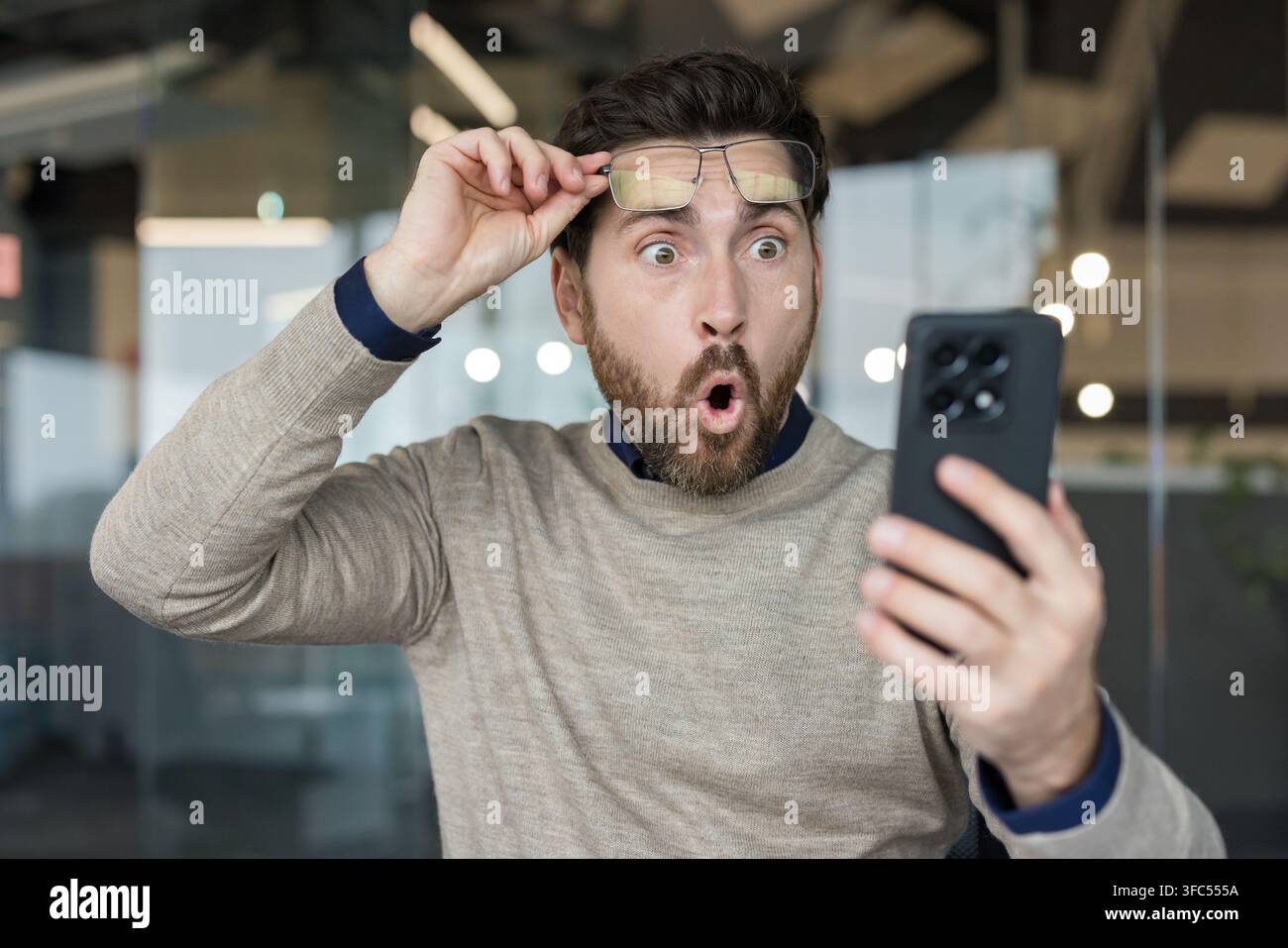 Uomo con gli occhiali spinti sulla fronte, che guarda uno smartphone con un'espressione di shock e incredulità, occhi spalancati e bocca aperta, mentre riceve notizie o contenuti inaspettati Foto Stock