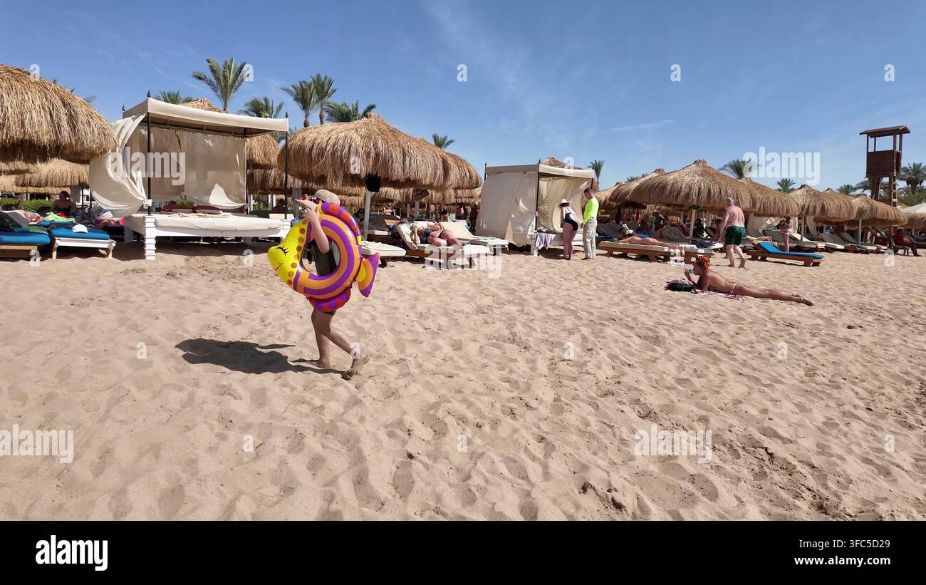 SHARM EL SHEIKH, EGITTO - 10 GIUGNO 2026: Un ospite che trasporta un giocattolo gonfiabile a forma di pesce sulla sabbia di un resort balneare in Egitto. Foto Stock