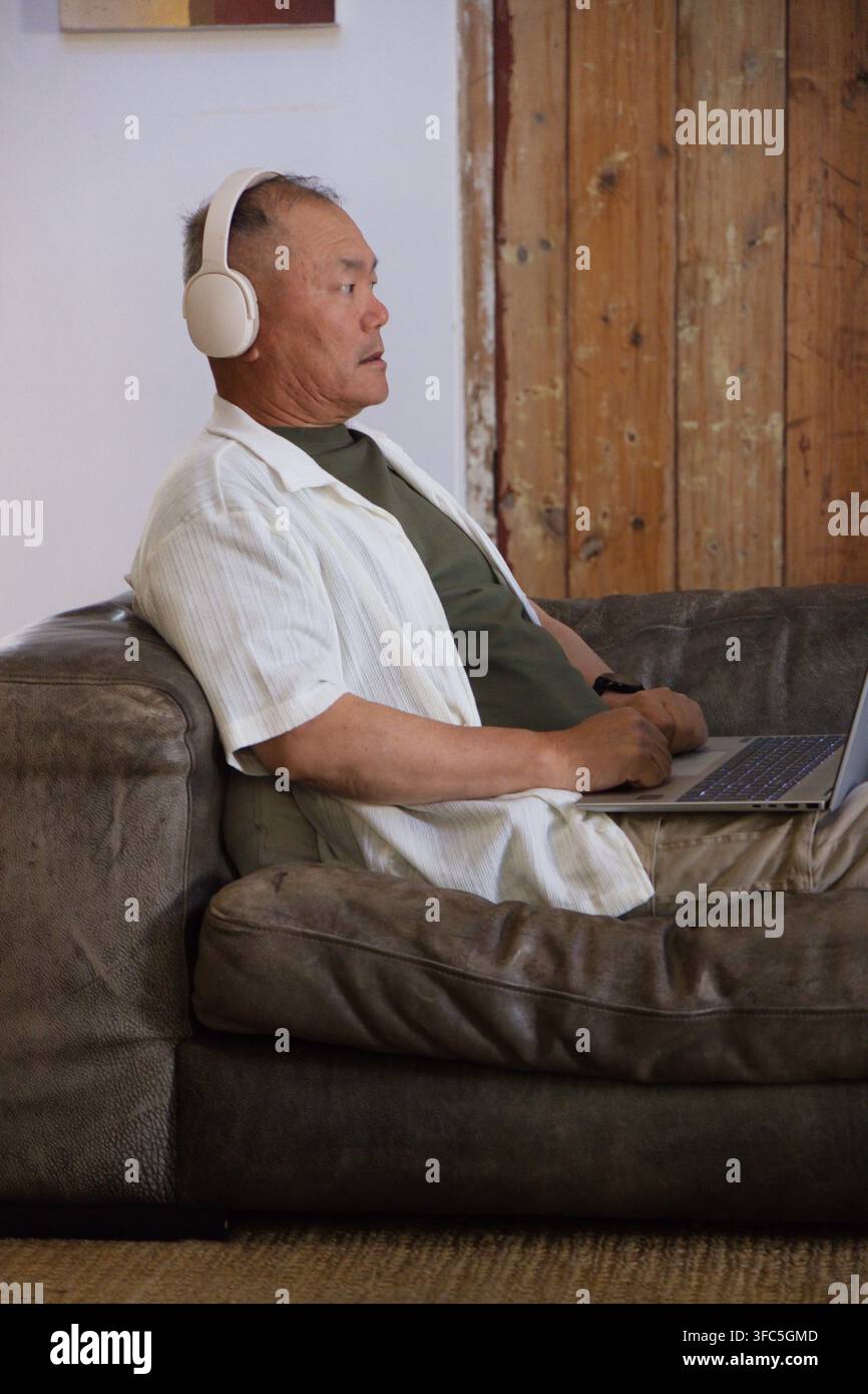 Uomo anziano asiatico seduto su un divano di pelle marrone scuro, con le cuffie e che digita su un laptop Foto Stock