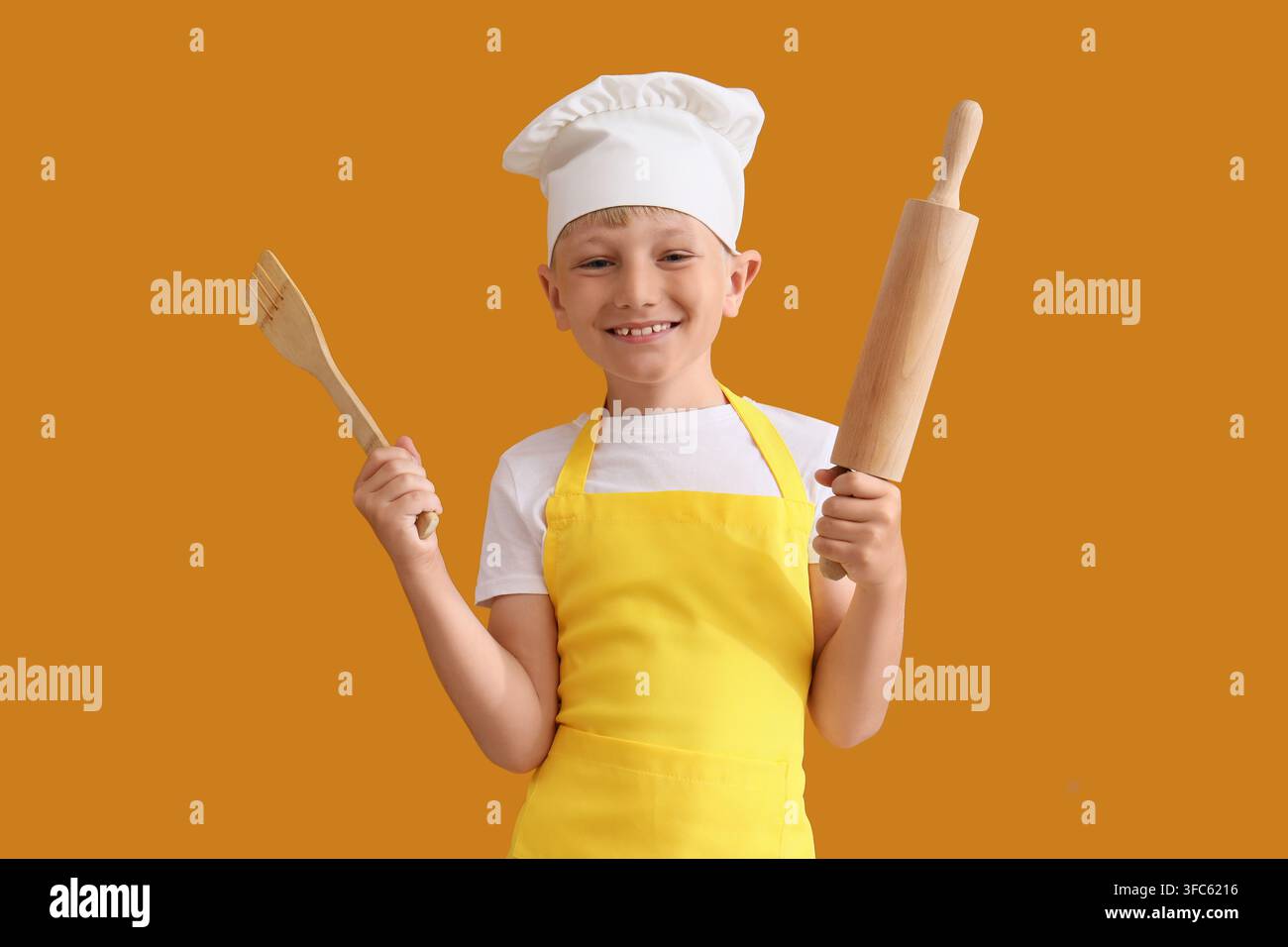 Concetto di chef bambino, ragazzo felice con grembiule e cappello che tiene utensili da cucina su sfondo arancione Foto Stock