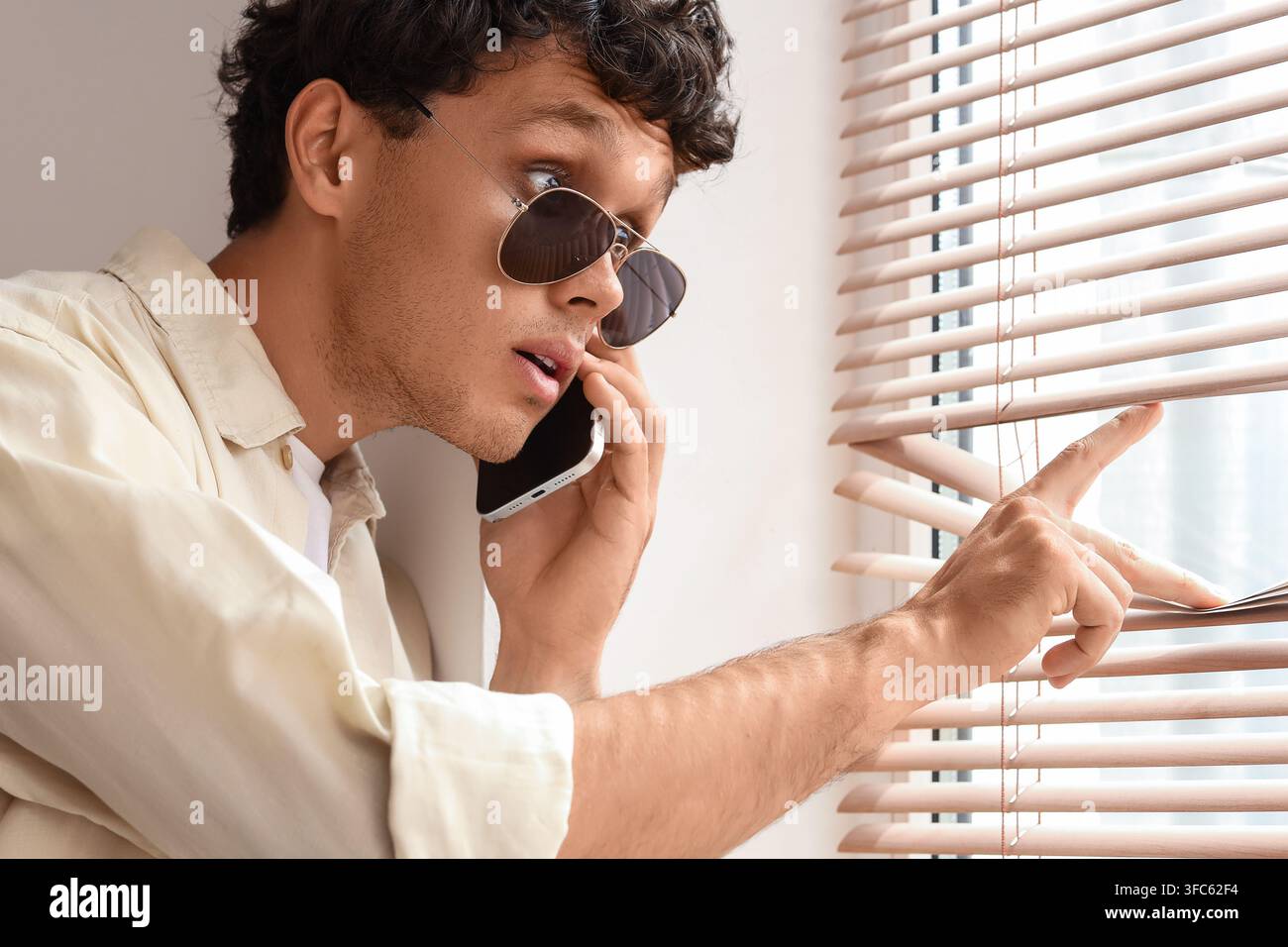 Sorveglianza e invasione della privacy, uomo con occhiali da sole al telefono mentre sbircia attraverso le persiane, spionaggio e indagine Foto Stock