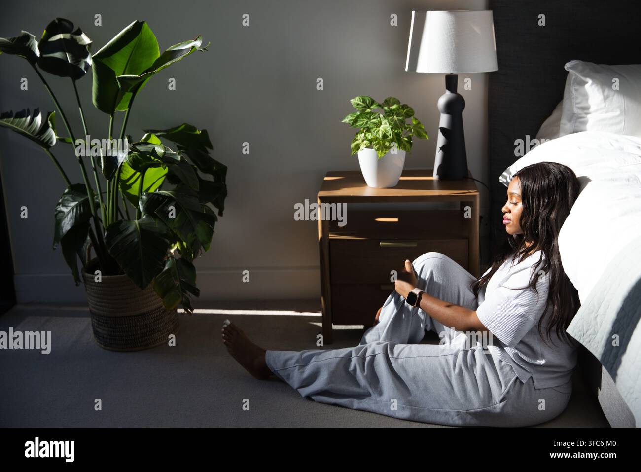 Donna africana seduta accanto al letto su un tappeto, indossa abbigliamento da relax grigio, smartwatch, pianta, spazio di copia Foto Stock