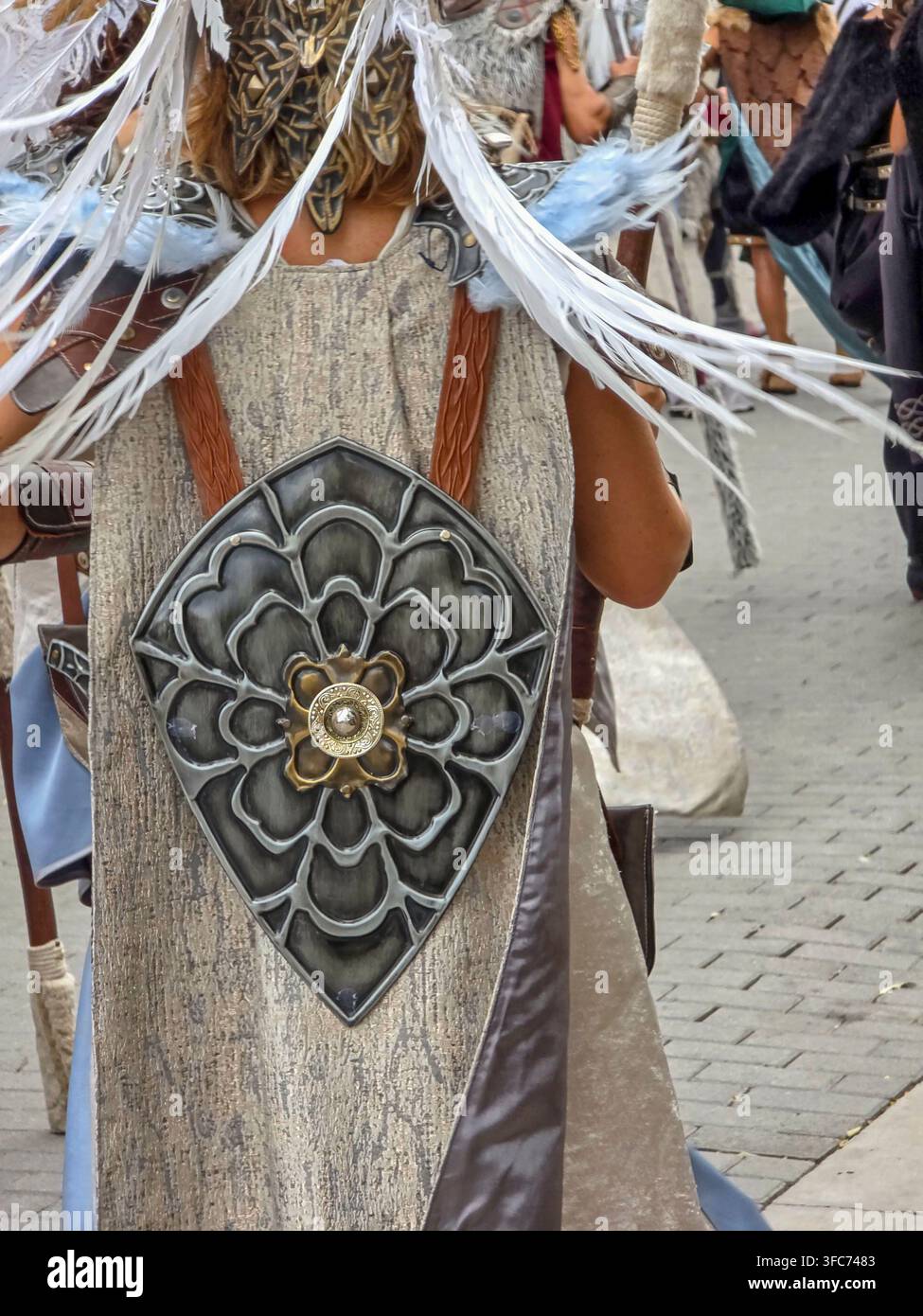 Una persona partecipa a un festival indossando un mantello grigio strutturato. Uno scudo decorativo metallico a forma di fiore con piume è attaccato alla schiena. P Foto Stock