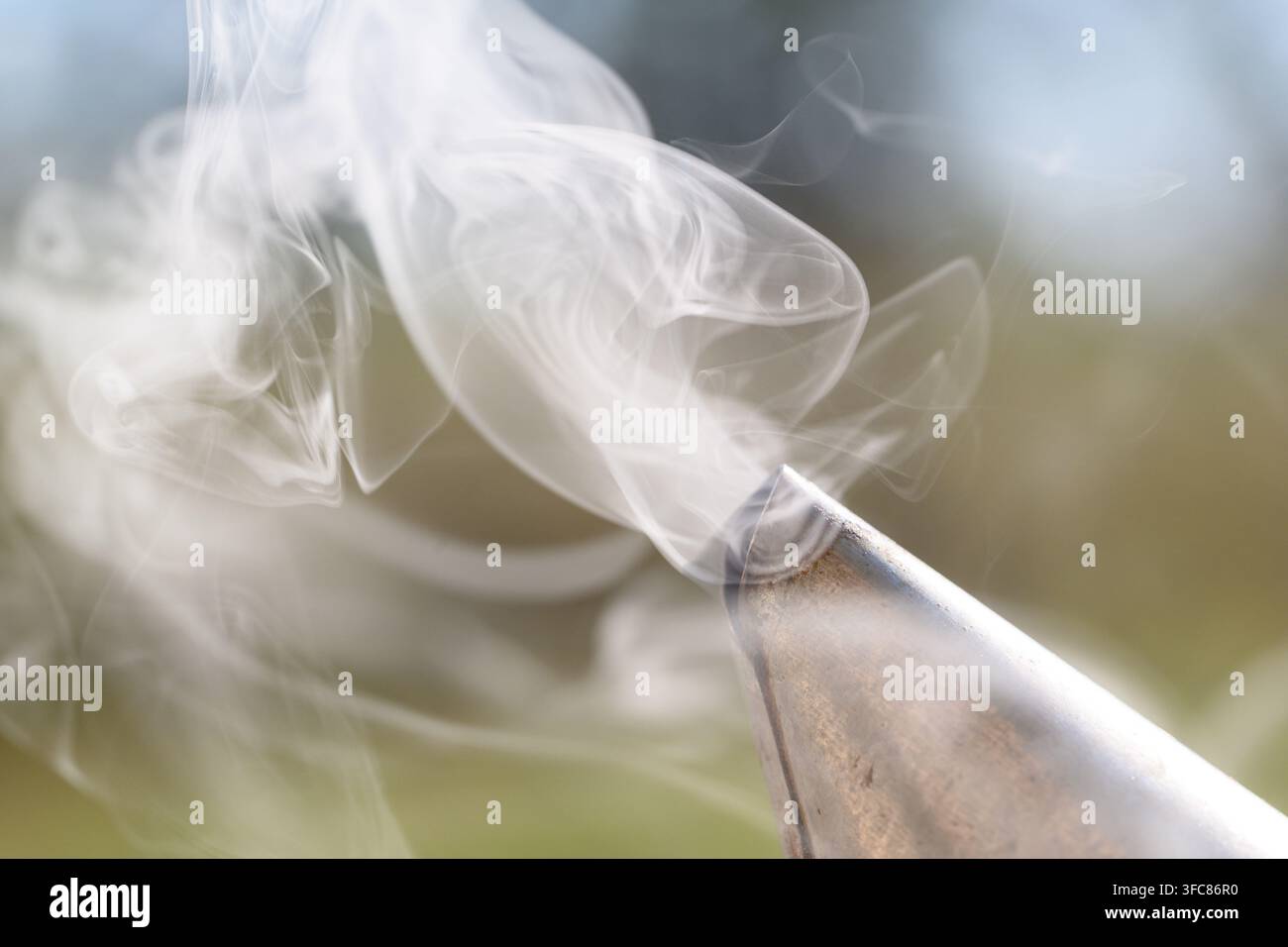 Fumo denso e arricciato si alza da un beccuccio per fumare le api, creando intricati motivi a vortice su uno sfondo verde sfocato. Foto Stock
