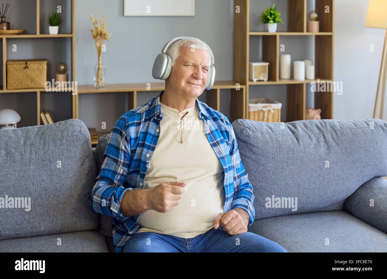 Uomo anziano soddisfatto che ascolta musica a casa sul divano con le cuffie comodamente Foto Stock