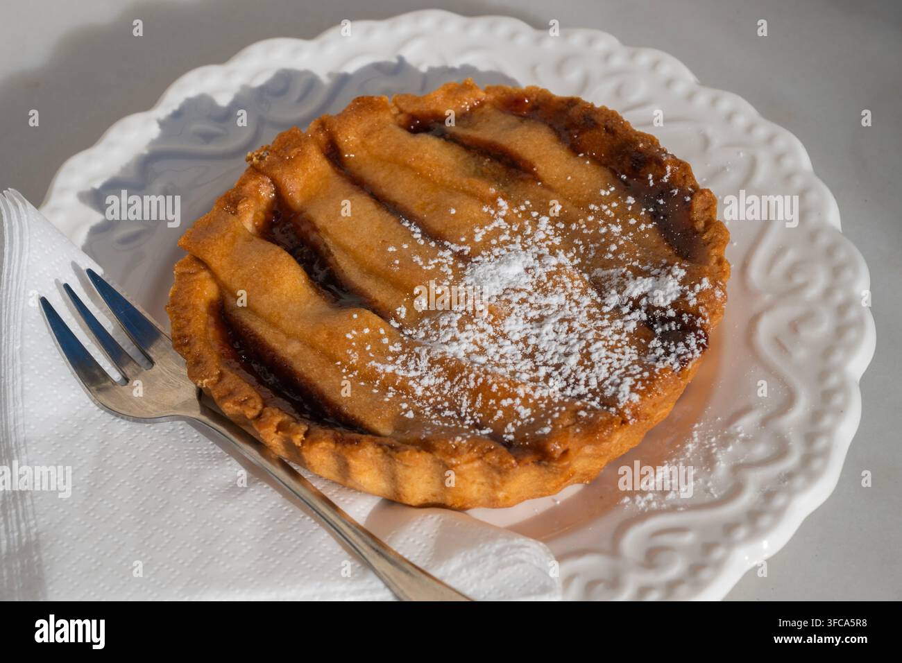 Una mini crostata ai frutti appena sfornata con una crosta a graticola dorata, spolverata di zucchero a velo, è servita su un piatto di ceramica bianco ornato con un fo Foto Stock