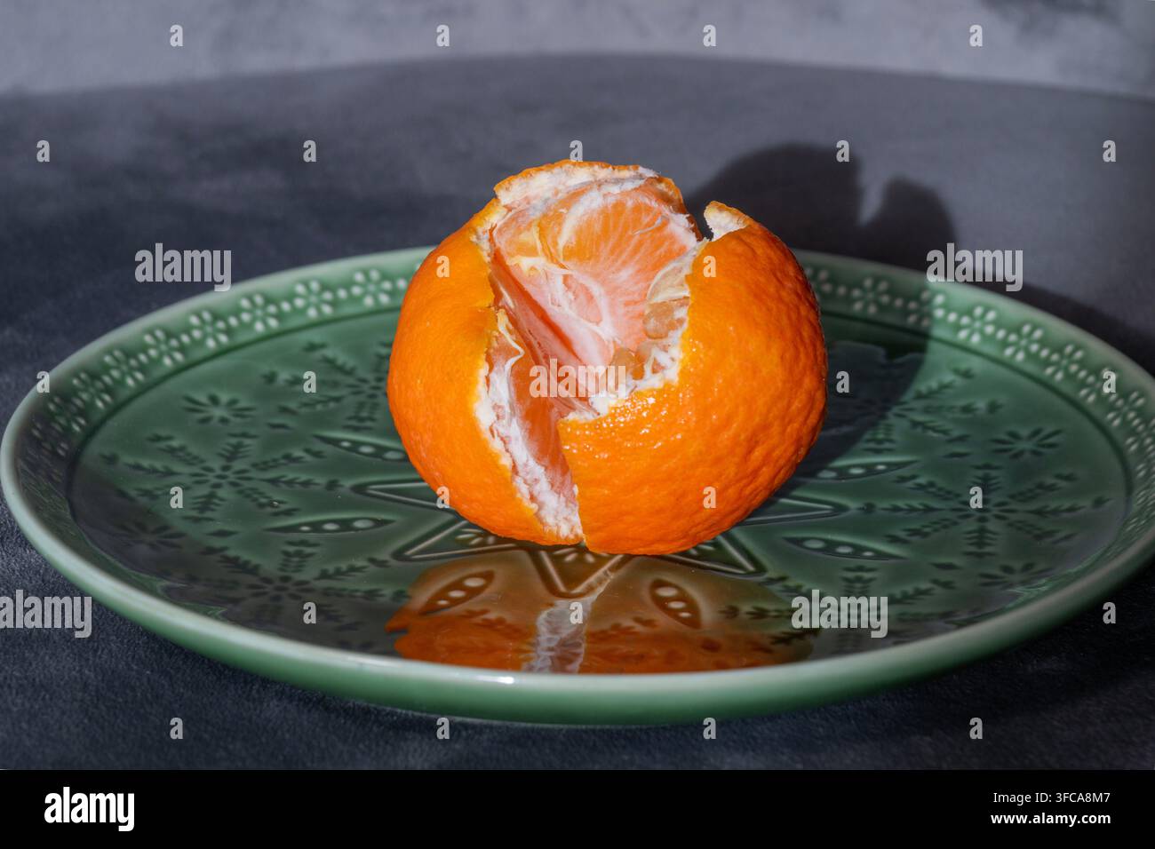 Una mandarina fresca parzialmente sbucciata è centrata su un piatto ornato con motivi verdi su uno sfondo scuro. Foto Stock
