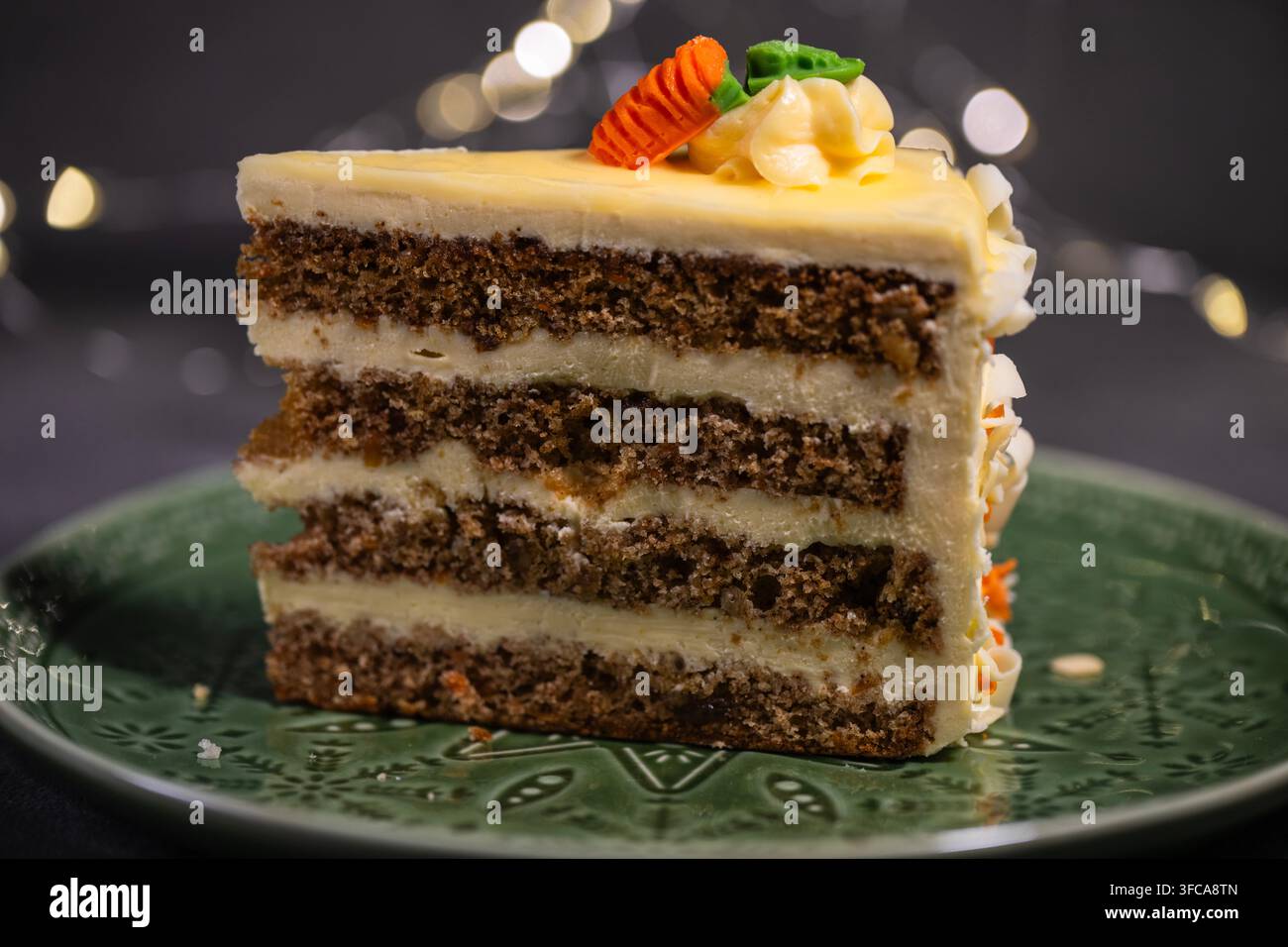 Una fetta di torta alle carote multistrato con glassa al formaggio cremoso, decorata con una piccola carota di zucchero, è posata su un piatto verde a motivi. Foto Stock