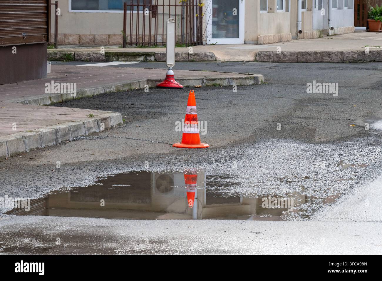 Un grande buca riempita d'acqua su una strada urbana è segnalata con un cono di segnalazione arancione brillante. Foto Stock