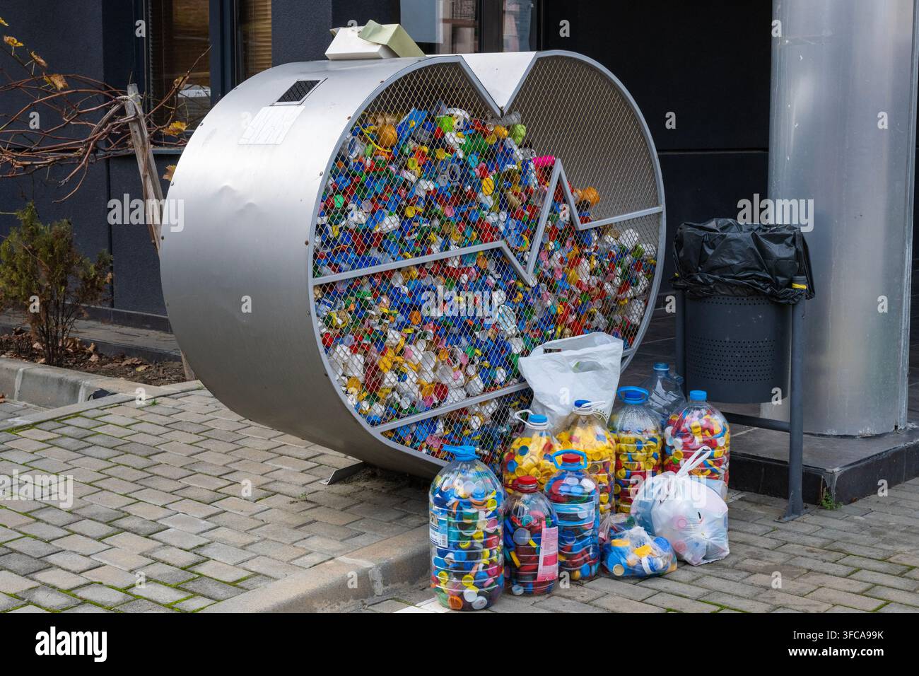 Un contenitore di riciclo a maglie metalliche a forma di cuore e bidoni di plastica circostanti traboccano di tappi di bottiglia di plastica colorati su un marciapiede urbano Foto Stock