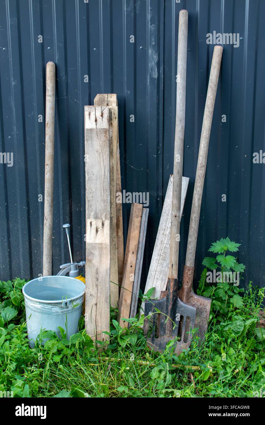 Pale da giardino e pali di legno appoggiati a un muro di metallo scuro Foto Stock