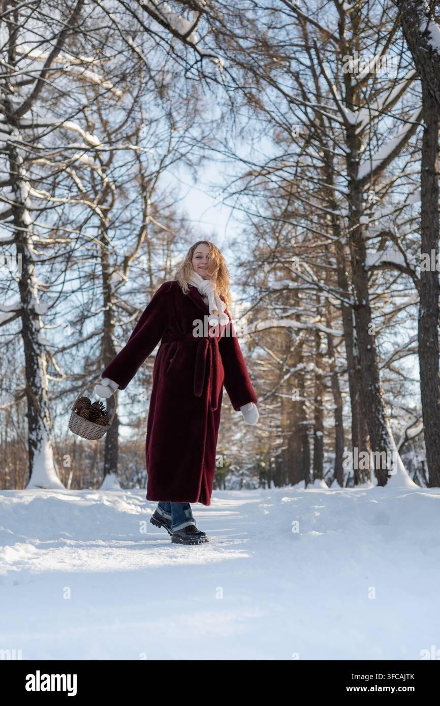 Donna in cappotto bordeaux cammina con un cestino di pigne lungo un sentiero del parco innevato in una giornata invernale soleggiata, godendosi la natura e lo stile di vita stagionale all'aperto. Vert Foto Stock