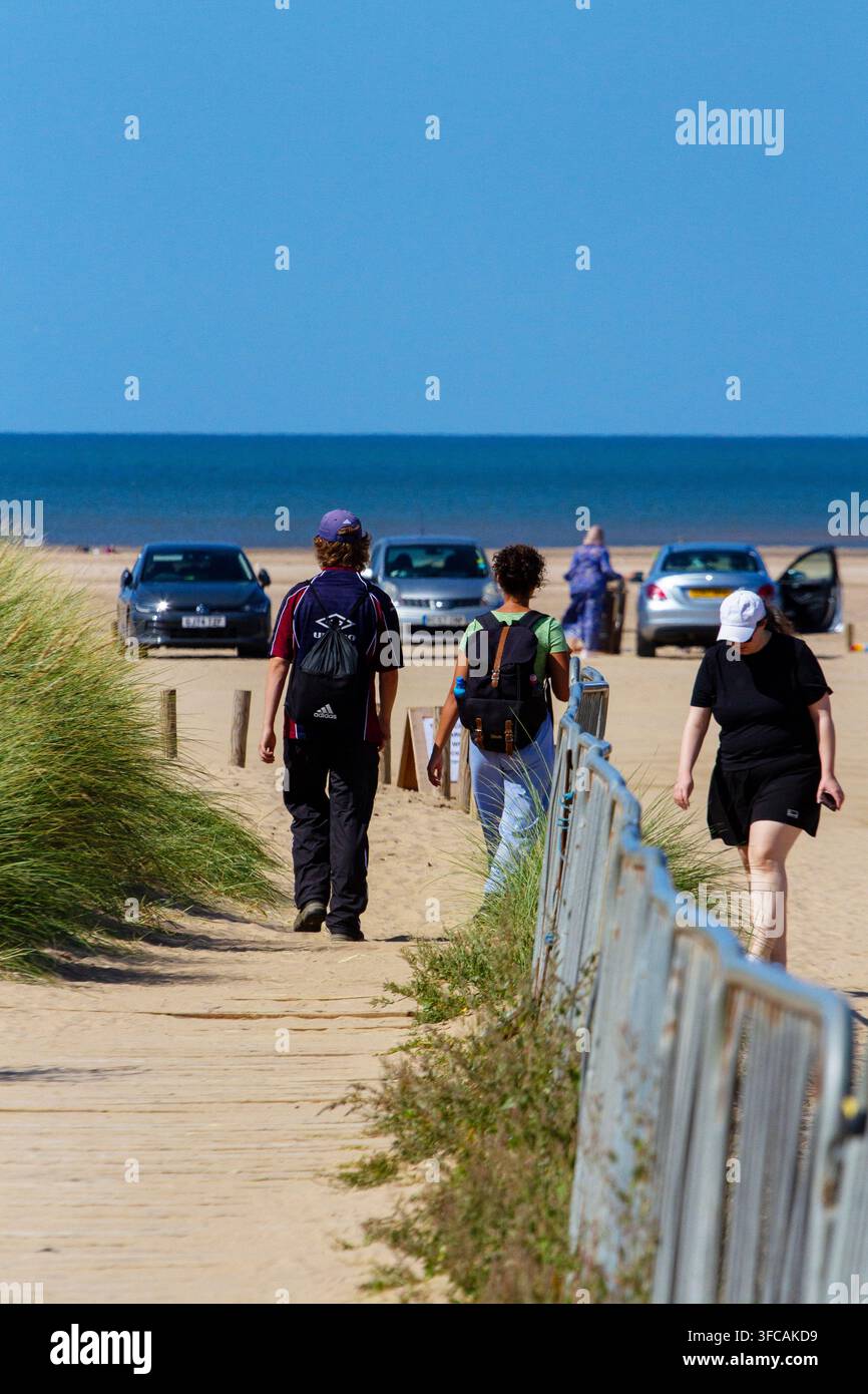 Gli escursionisti con zaini passeggiano lungo un sentiero sabbioso dirigendosi verso le auto parcheggiate sulla spiaggia con l’oceano blu davanti. Southport, Regno Unito. Foto Stock