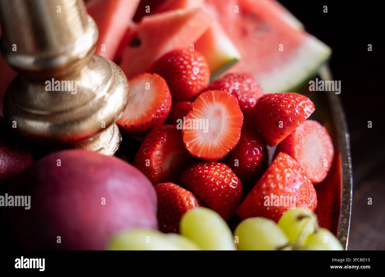 Macro primo piano di fragole rosse mature affettate circondate da fette di anguria e uva su un supporto decorativo dorato per servire Foto Stock