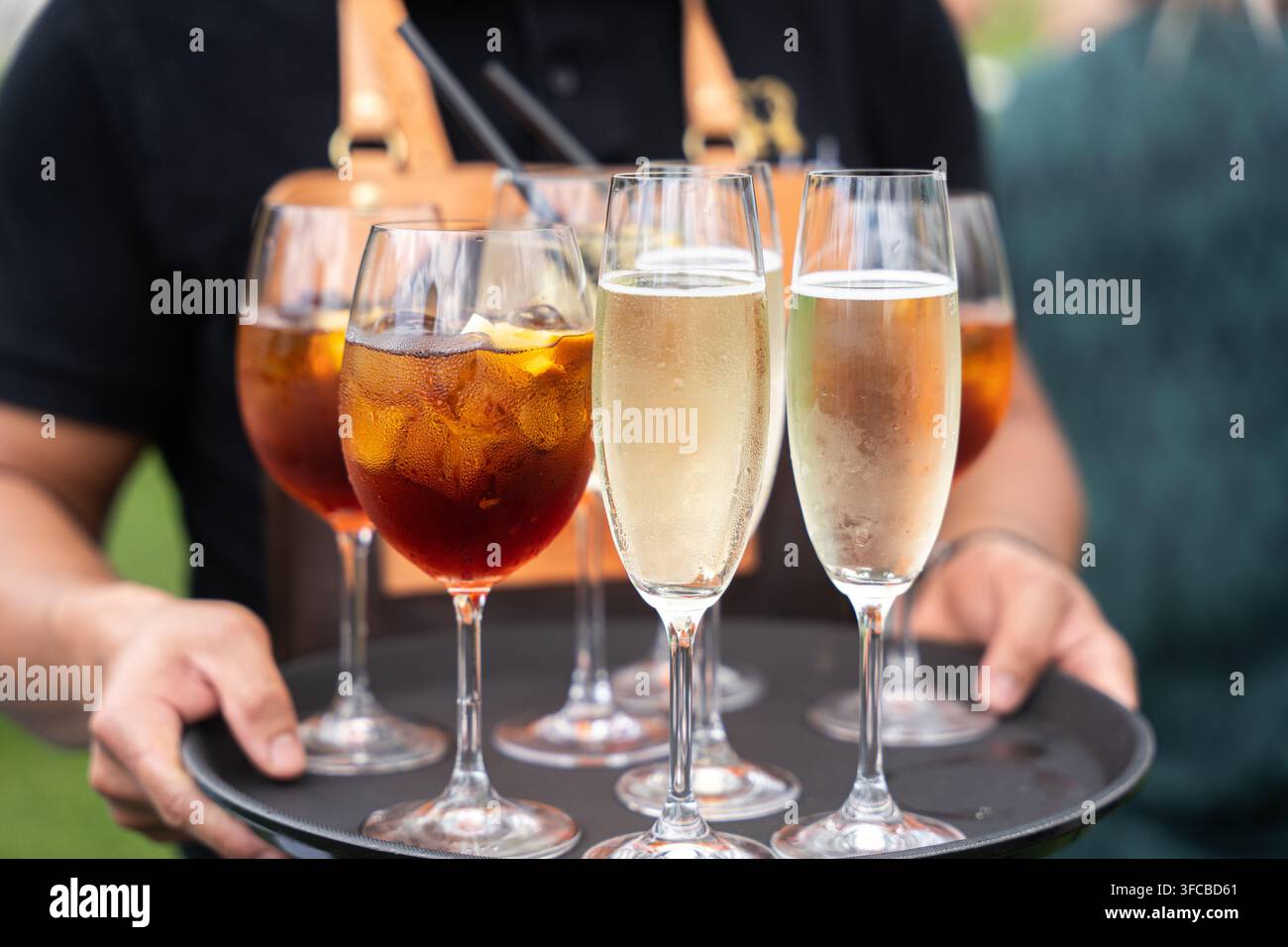 Cameriere professionista che serve più bicchieri di champagne raffreddato e cocktail vivaci su un vassoio nero, offrendo un servizio di ospitalità premium a un Foto Stock