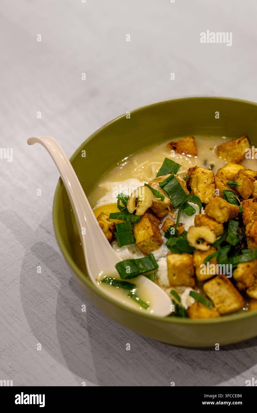 Zuppa calda con tofu croccante e verdure fresche. Ciotola verde contiene un pasto confortante e fumante. Cuchchiaio bianco riposa delicatamente accanto al cibo. Toni chiari suggeriscono Foto Stock