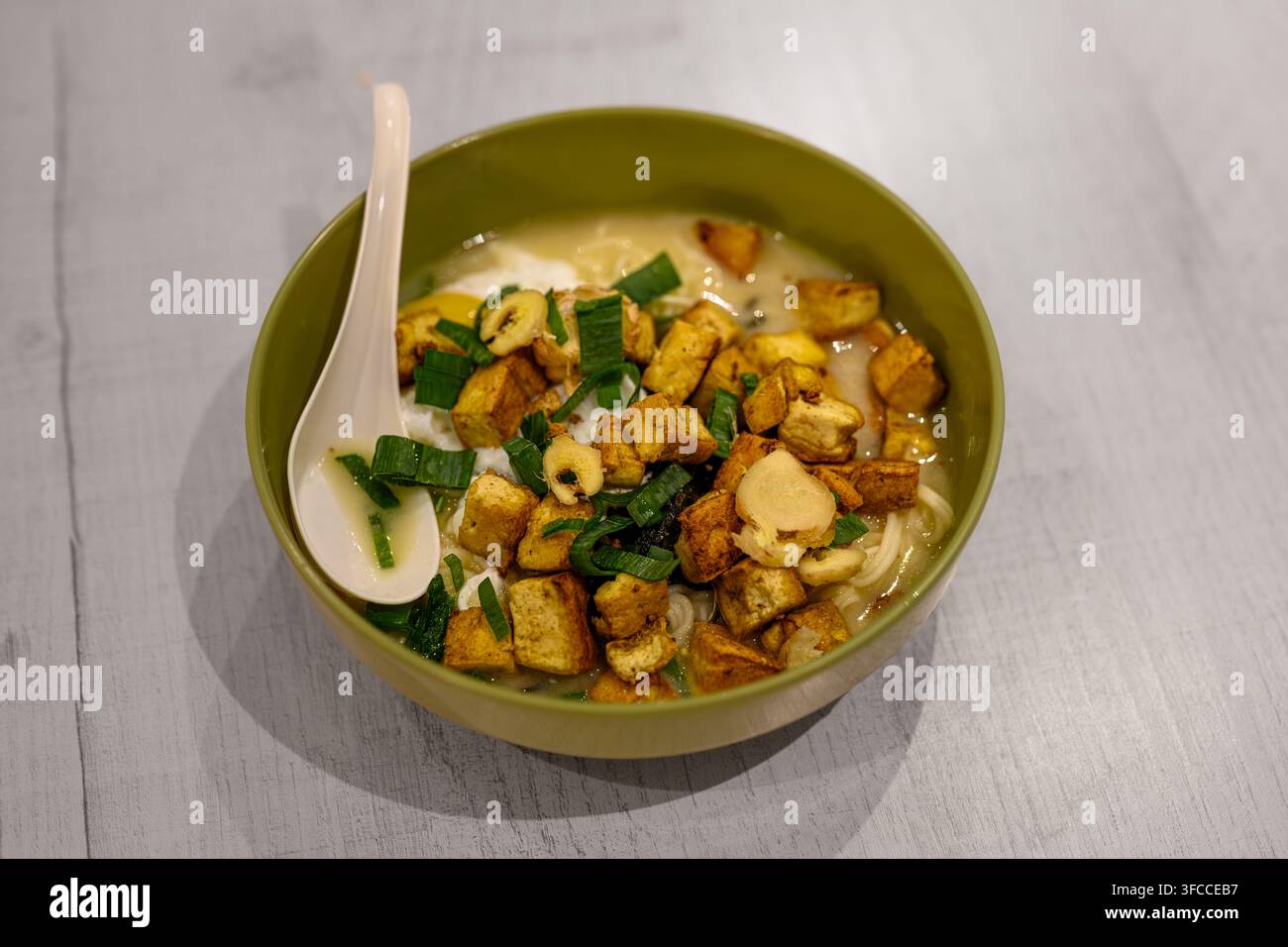 Zuppa calda con tofu croccante ed erbe fresche in una ciotola verde. Un cucchiaio bianco riposa accanto alla ciotola. Una superficie in legno chiaro aggiunge un fascino rustico. Ideale per cibo, com Foto Stock