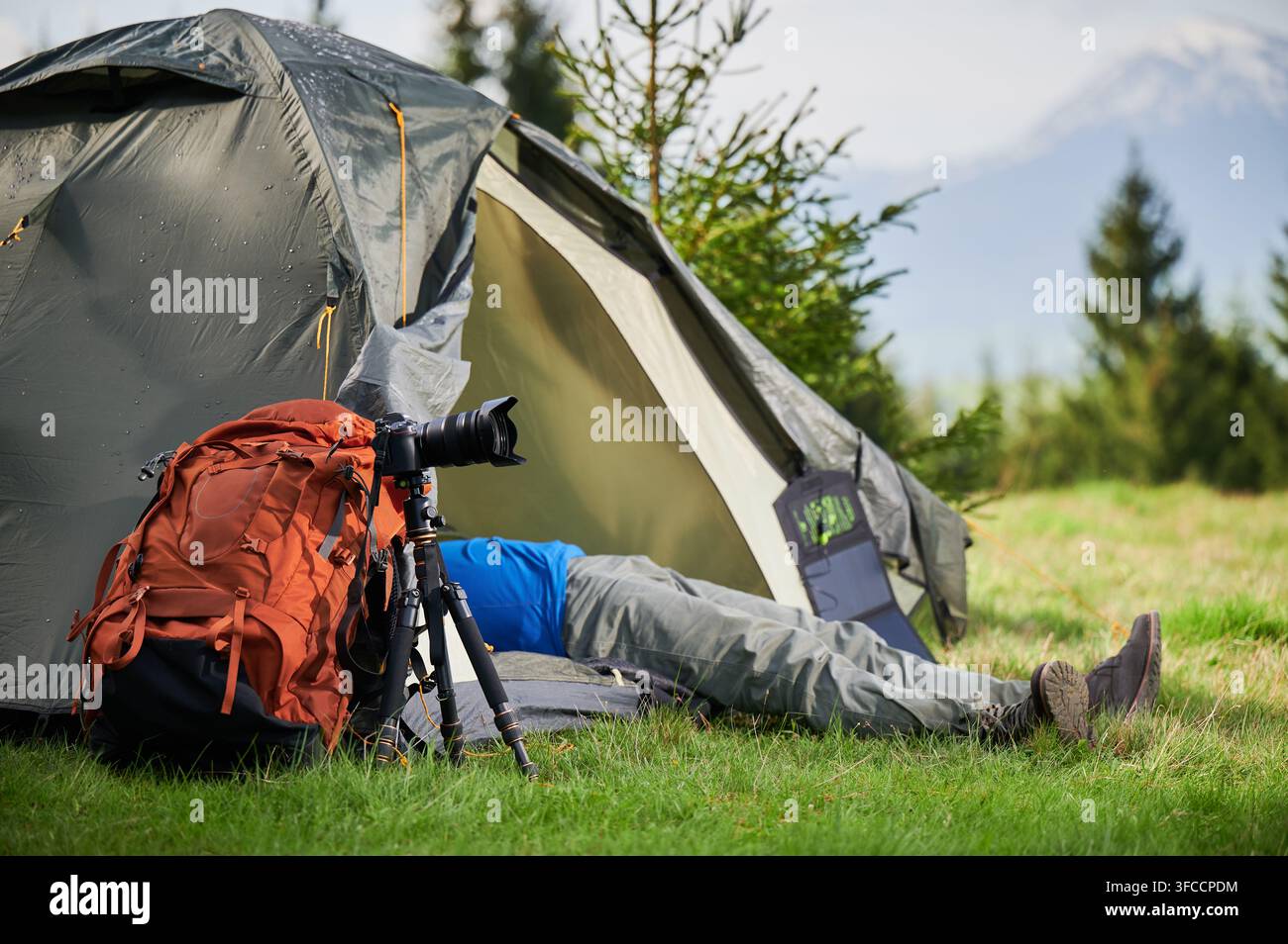 Fotografo di viaggio si riposa all'ingresso della tenda dopo una sessione fotografica all'aperto, con fotocamera, zaino e attrezzatura in un campeggio di montagna. Utile per il marketing avventuroso, la promozione del campeggio, concetti di stile di vita Foto Stock