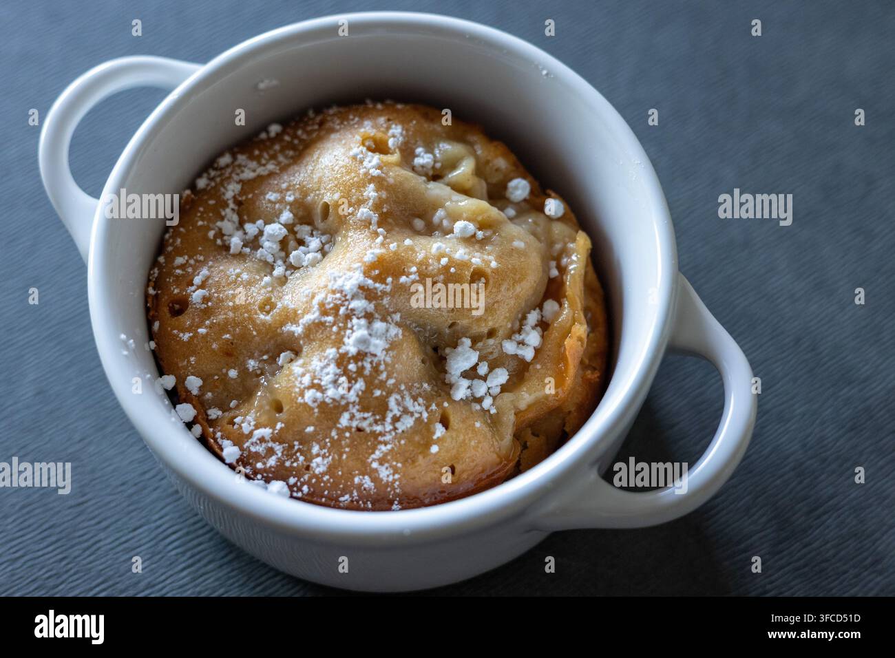 Vista angolata di una torta individuale alle mele dorata in una cocotte bianca a due manici, spolverata di zucchero a velo su un tessuto grigio strutturato Foto Stock