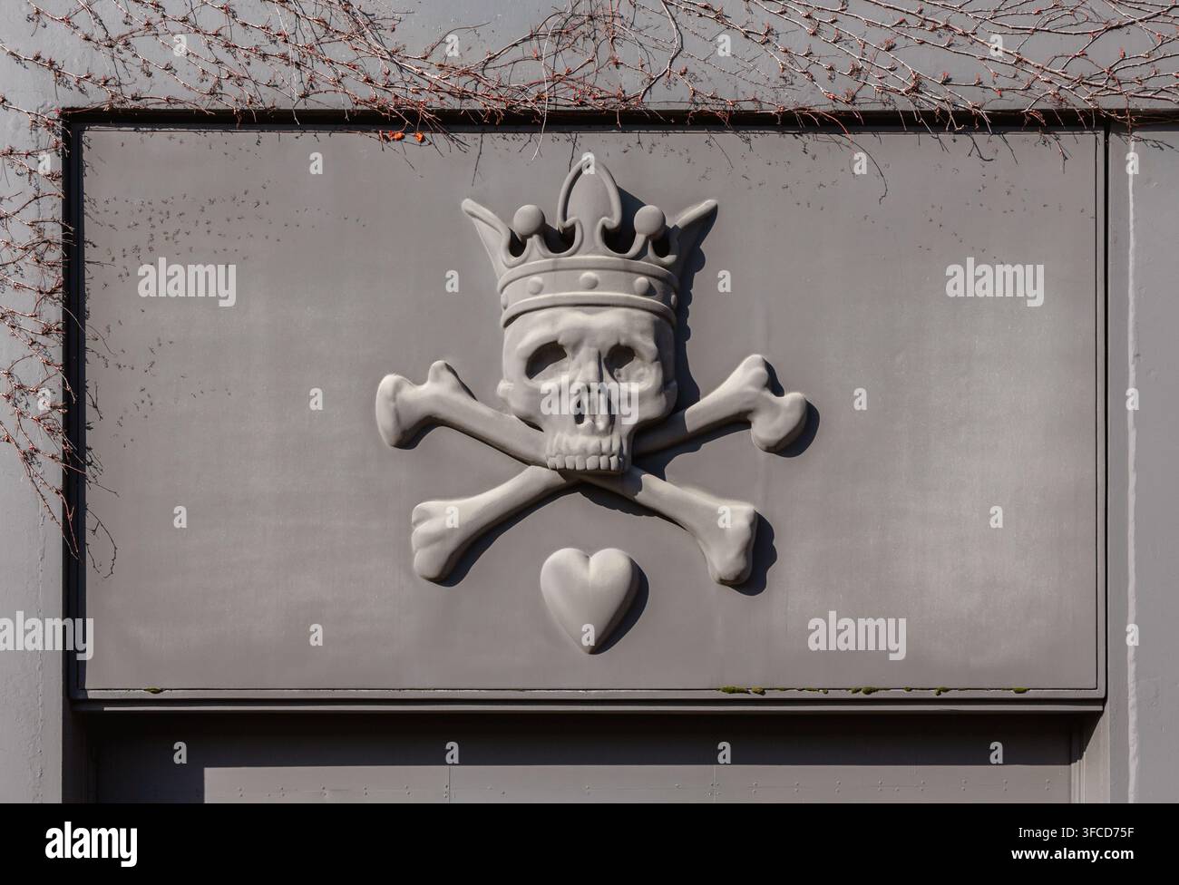 Scultura in rilievo in pietra di un teschio incoronato con ossa incrociate e un piccolo cuore montato su un muro che proietta un'ombra drammatica. Foto Stock