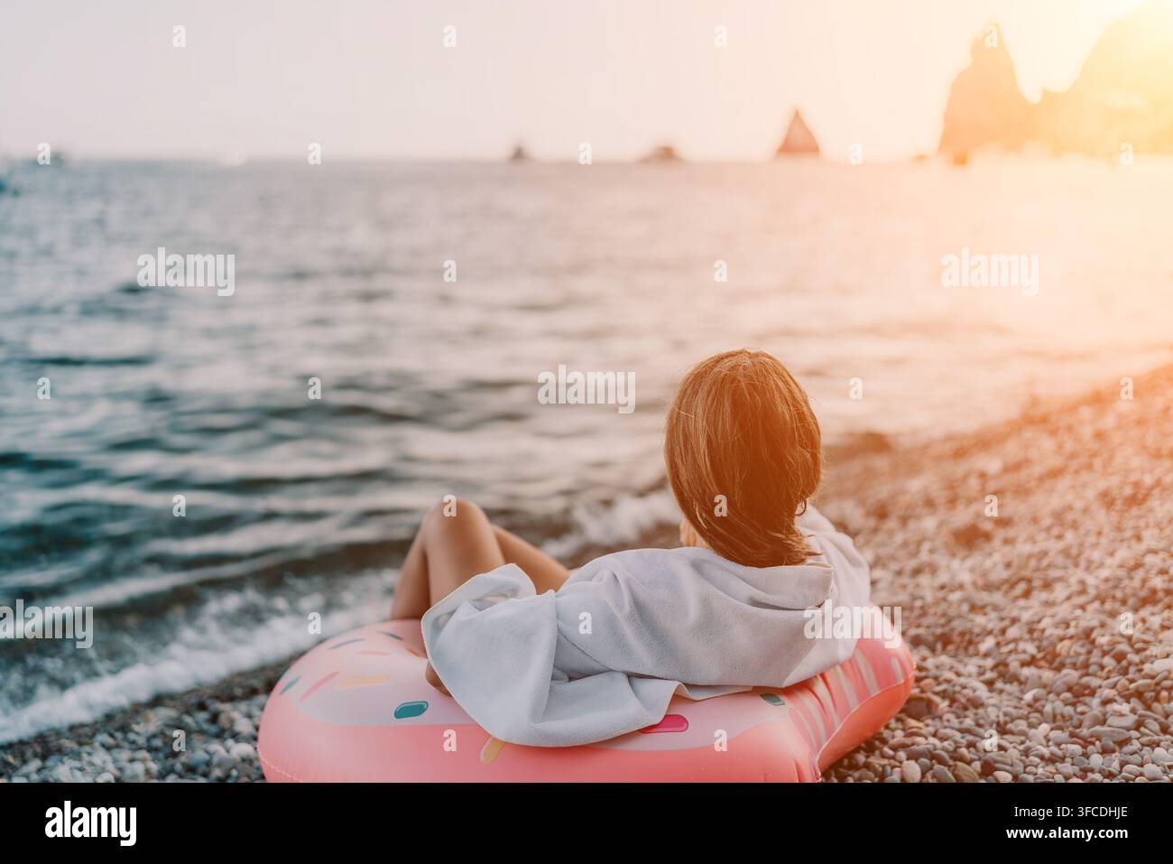 Rilassamento in vacanza al mare: donna si riposa su un galleggiante a forma di ciambella godendosi un tramonto tranquillo su una spiaggia di ciottoli. Foto Stock