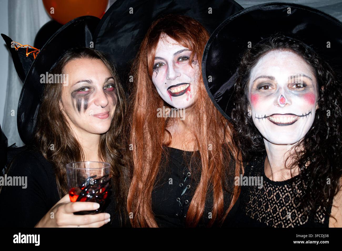 Tre donne che posano insieme con cappelli da strega e trucco di Halloween mentre si godono dei drink durante una festa in costume festosa. Foto Stock