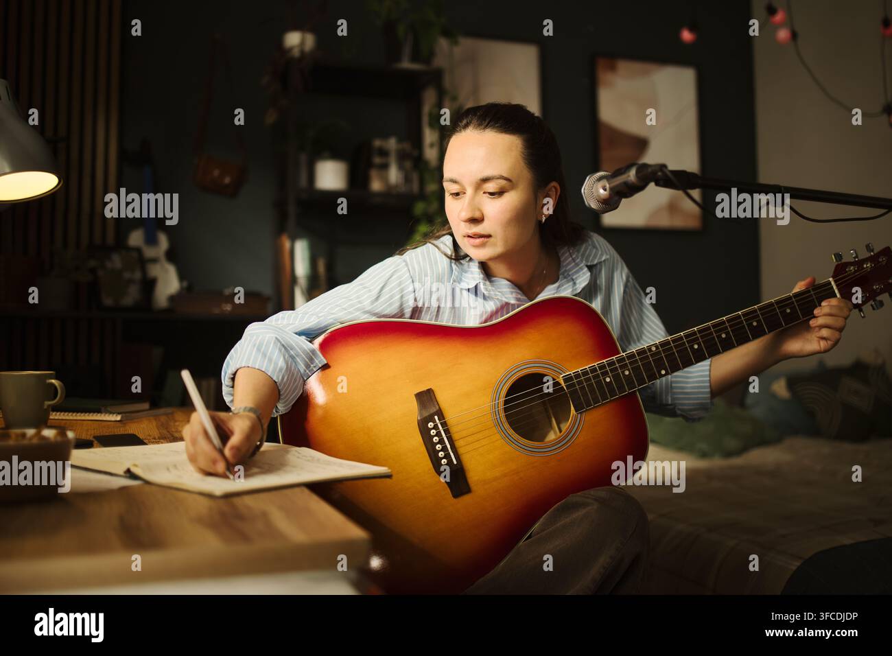 Giovane donna caucasica che tiene una chitarra acustica e scrive testi in un taccuino nello studio di casa. L'immagine rappresenta il processo creativo di scrittura di canzoni, adatto per il marketing dell'educazione musicale Foto Stock