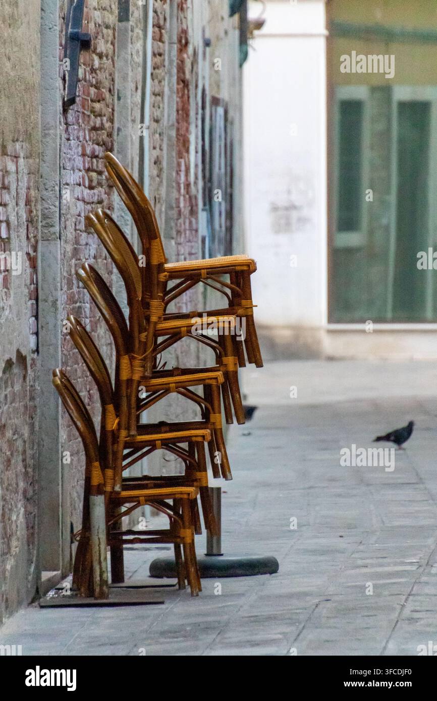 Sedie da caffè in legno impilate si appoggiano a un vecchio muro di mattoni in una stretta strada tranquilla, con un piccione che cammina sul marciapiede. Foto Stock