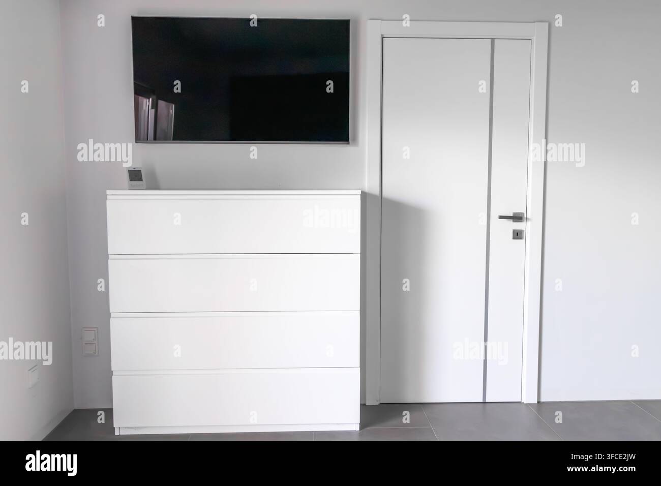 Un interno di camera da letto minimalista con un elegante cassettone bianco con cassetti e una televisione a parete sopra di esso Foto Stock