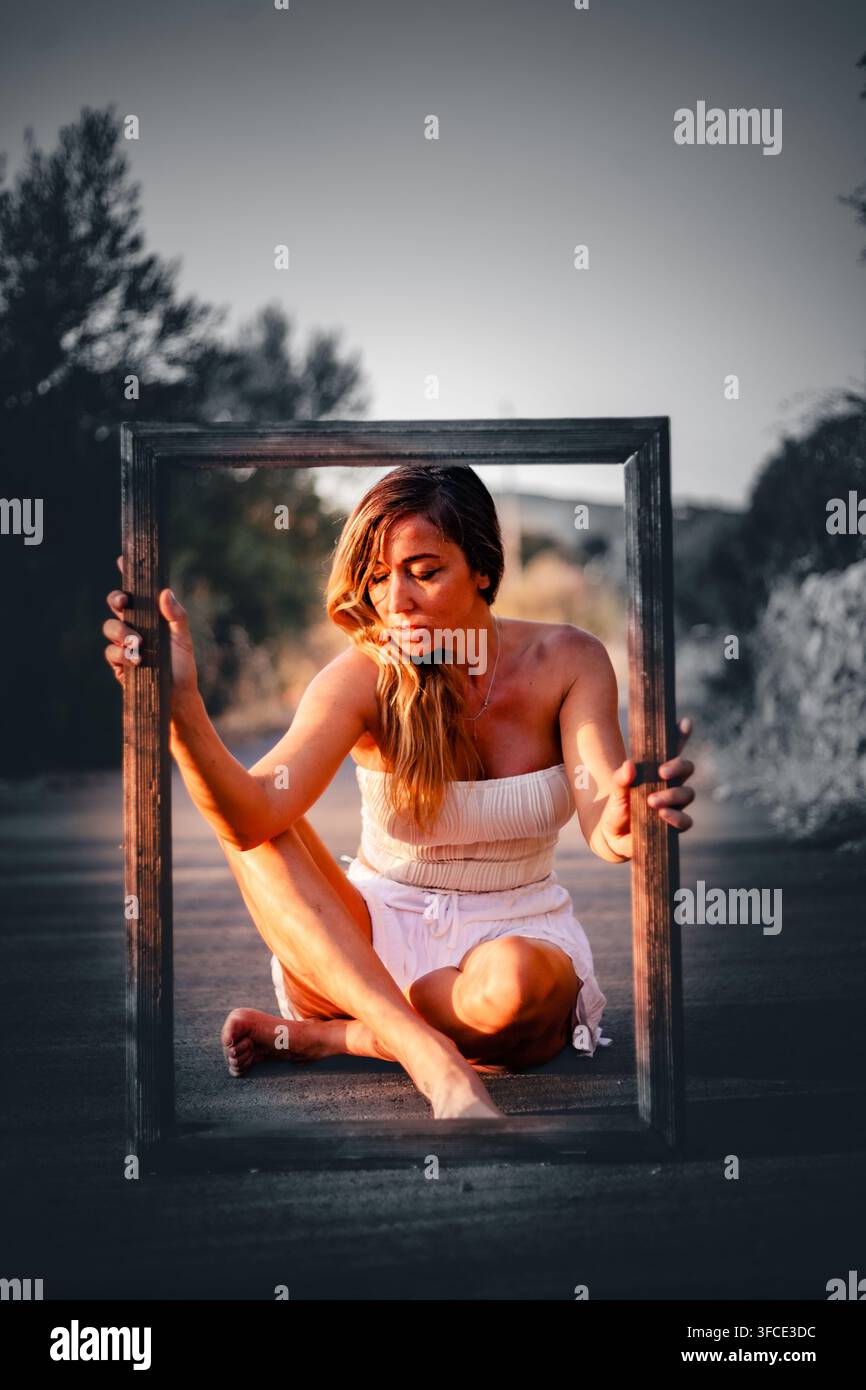 Giovane donna in abito bianco seduta a piedi nudi su una strada rurale tenendo una grande cornice fotografica in legno al tramonto, Algarve, Portogallo Foto Stock