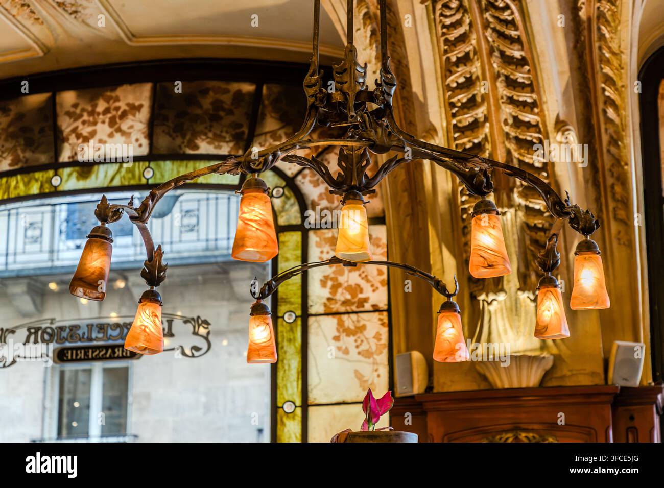 Un lampadario Art Nouveau con diversi paralumi in vetro arancione è sospeso davanti a una finestra in vetro colorato alla Brasserie Excelsior di Nancy, Grand Est, Francia, attraverso la quale si può leggere la scritta "L'Excelsior Brasserie" specchiata. Foto Stock