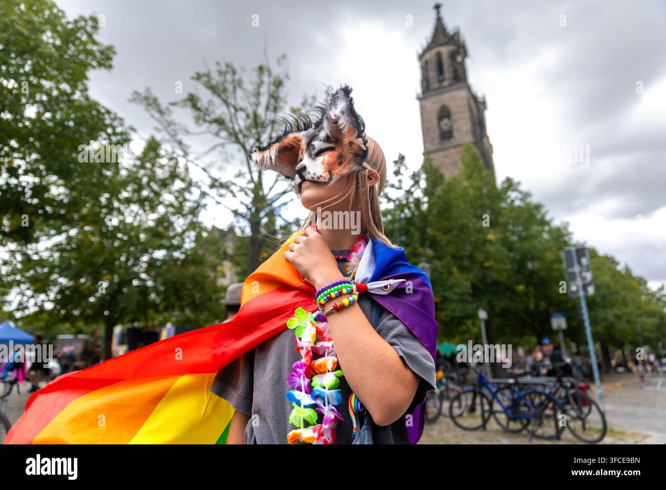 Ragazza theriana mascherata in mantello arcobaleno colorato posa durante la celebrazione del CSD a Magdeburgo, Germania, con la cattedrale sullo sfondo. La scena riflette la gioventù Foto Stock