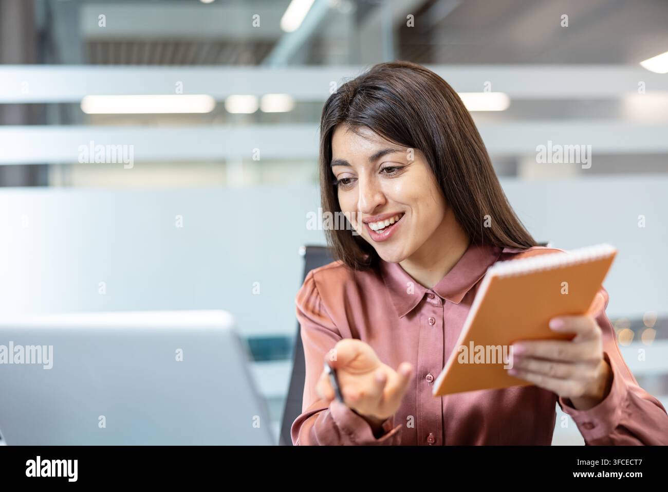 Giovane donna professionista che tiene una allegra videochiamata su un laptop, discute di lavoro e prende appunti in un quaderno durante una riunione remota in un ambiente di ufficio moderno Foto Stock
