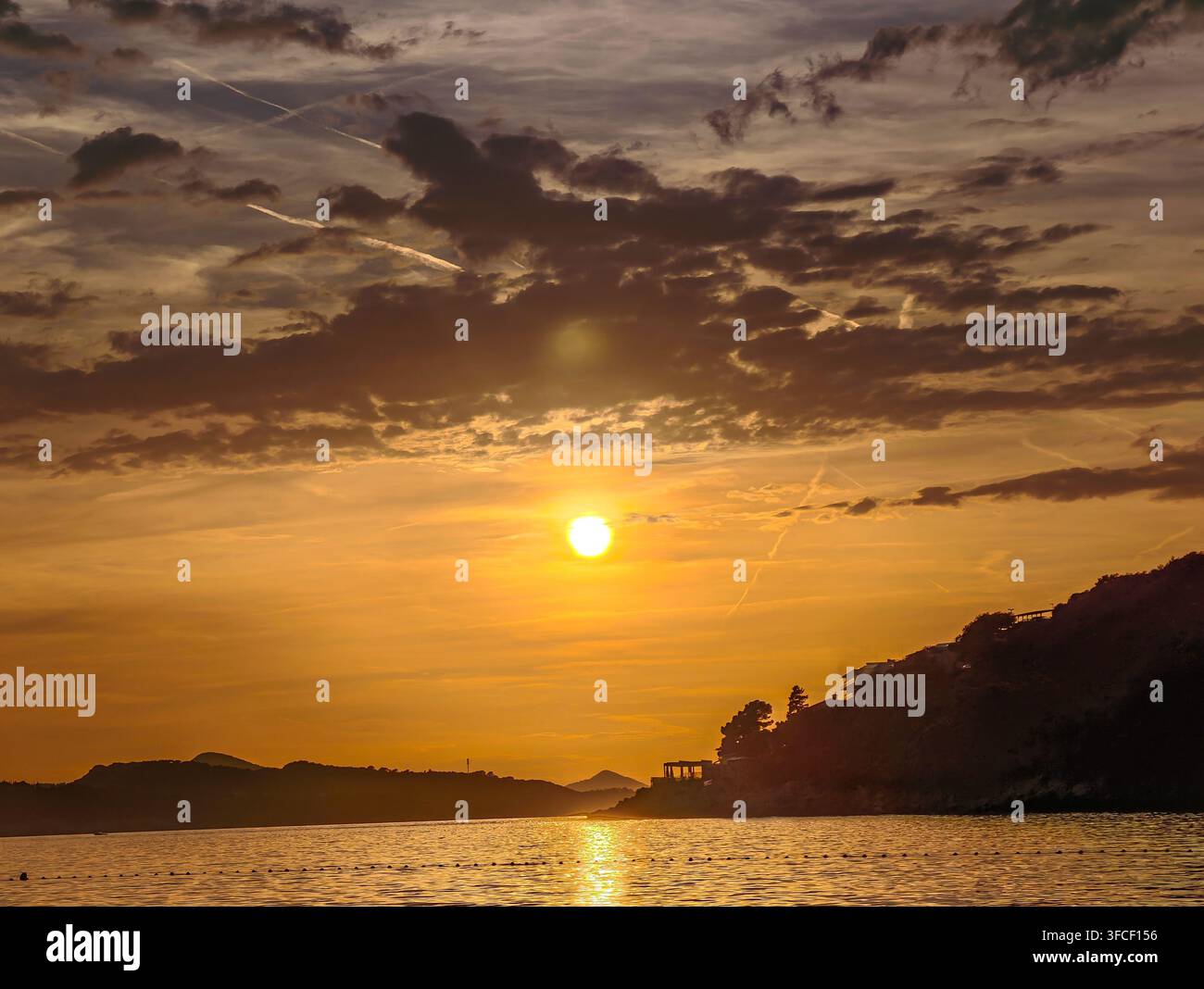Un tramonto dorato mozzafiato dipinge il cielo sopra le acque calme dell'oceano, proiettando silhouette sulla costa frastagliata in un momento di pura pace. - Immagine stock catturata con smartphone