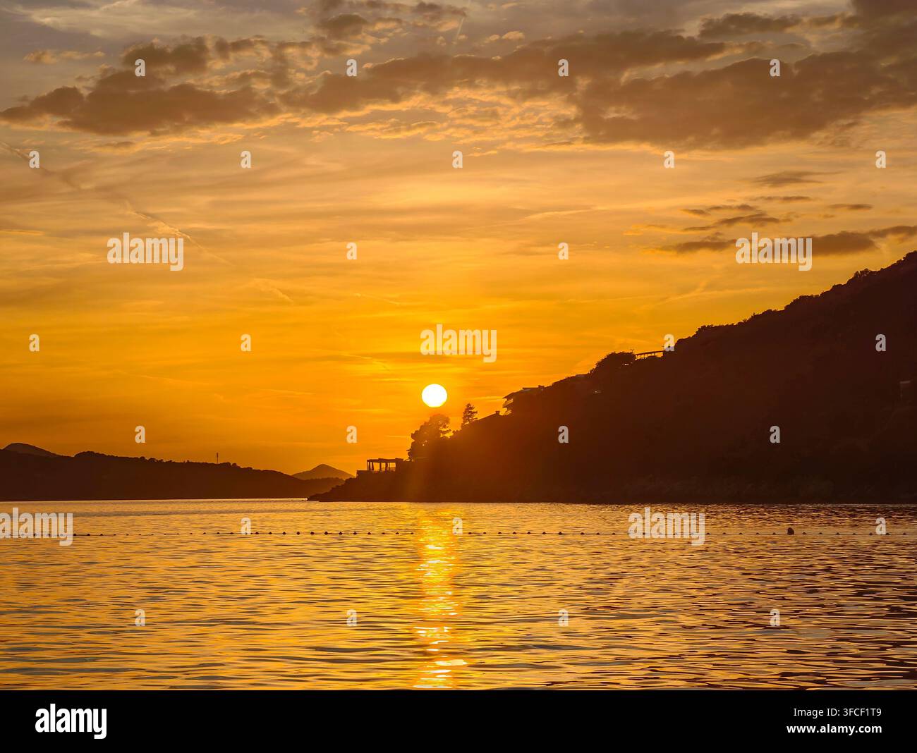 Un tramonto dorato mozzafiato dipinge il cielo sopra le acque calme dell'oceano, proiettando silhouette sulla costa frastagliata in un momento di pura pace. - Immagine stock catturata con smartphone