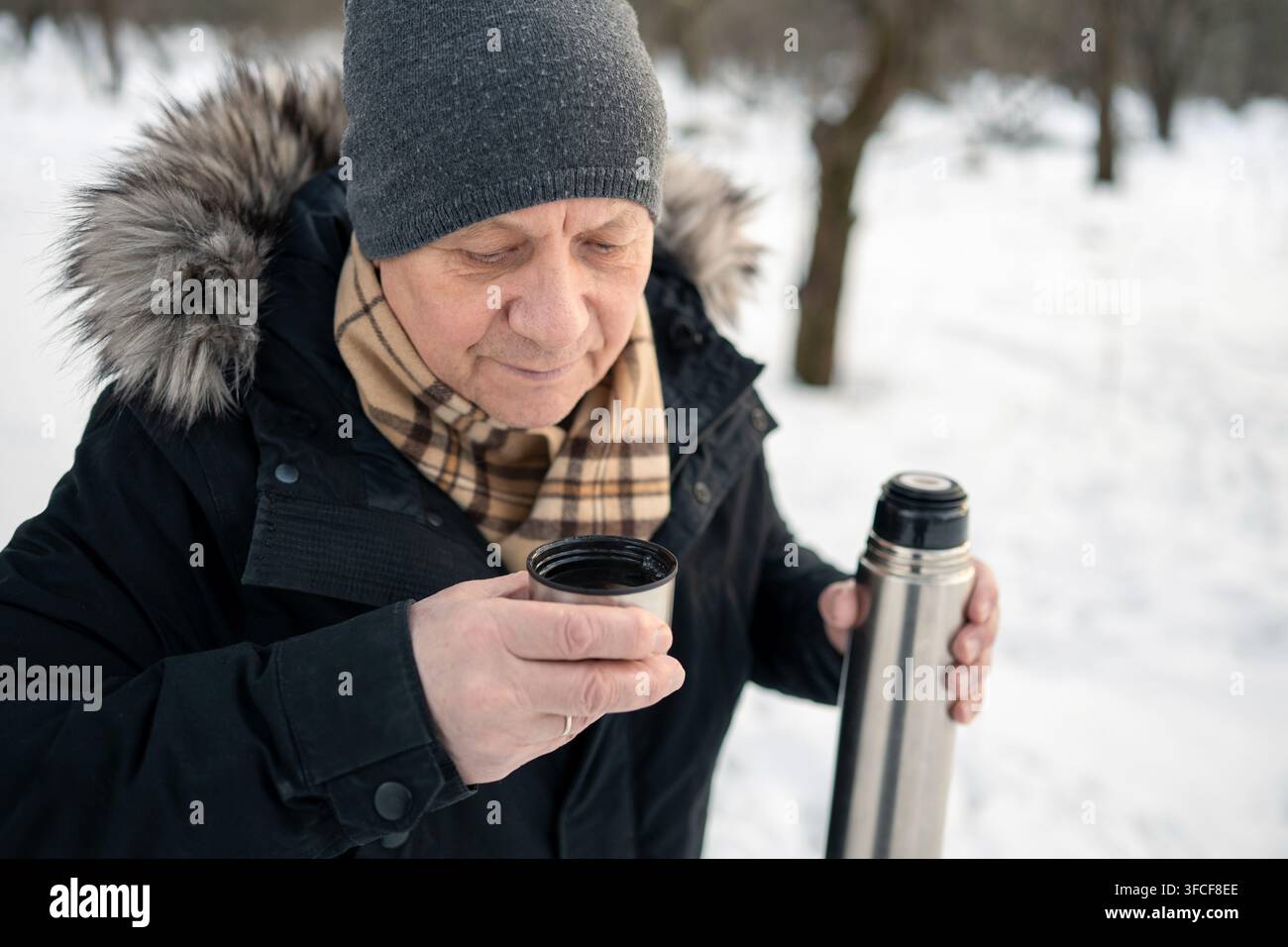 Uomo anziano che tiene una thermos e una tazza con una bevanda calda all'aperto in un parco innevato. Bevanda riscaldante durante una passeggiata invernale, invecchiamento attivo e stile di vita con il freddo Foto Stock