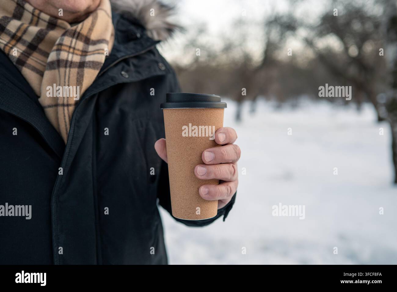Uomo anziano che tiene una tazza da asporto riutilizzabile per il caffè durante una passeggiata invernale in un parco innevato. Bevanda calda, stile di vita nel freddo, invecchiamento attivo e svago all'aperto. Foto Stock