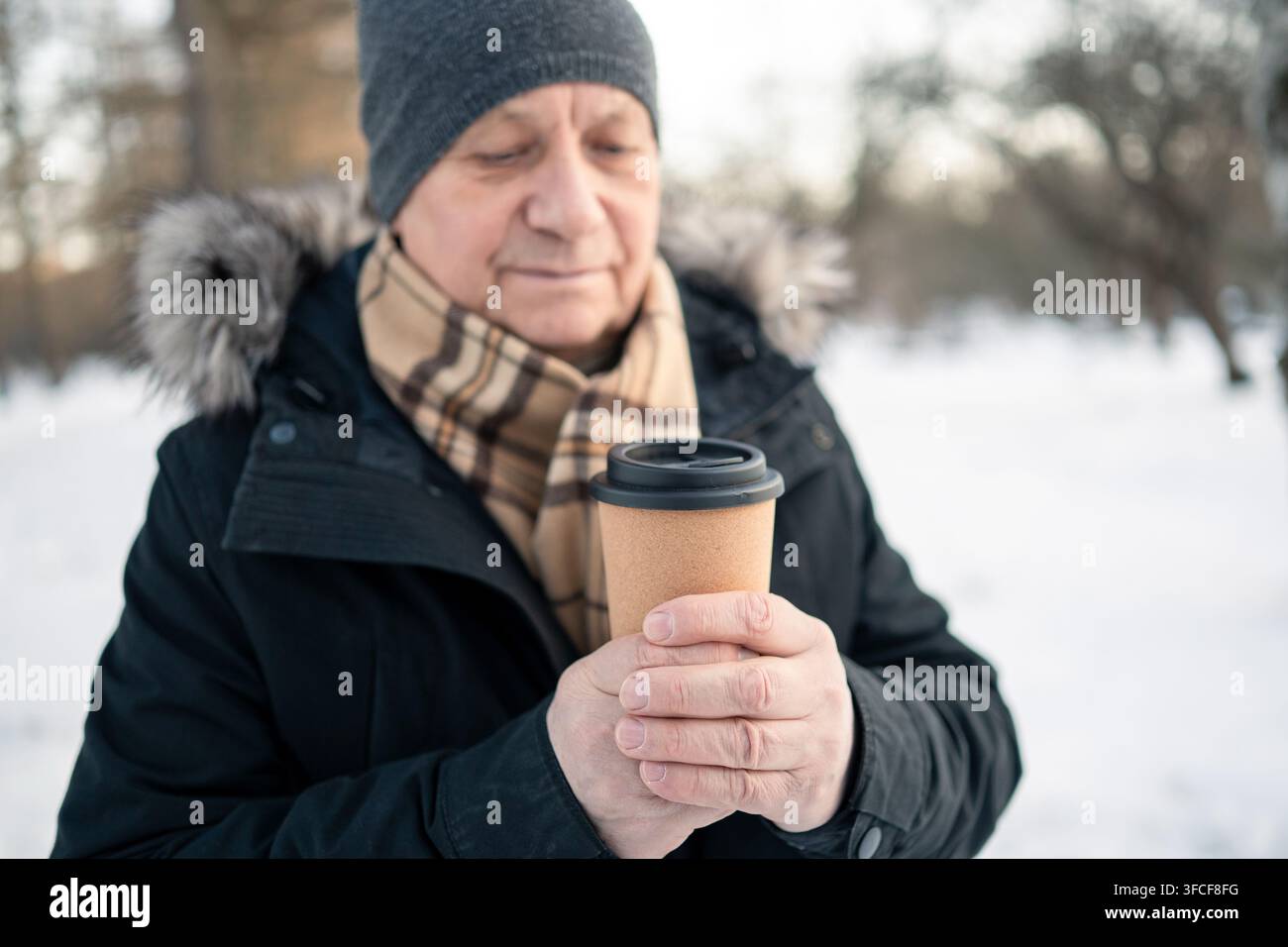 Uomo anziano che riscalda entrambe le mani attorno a una tazza di caffè da asporto all'aperto in un parco innevato. Bevanda calda, comfort invernale, freddo e stile di vita attivo degli anziani. Foto Stock