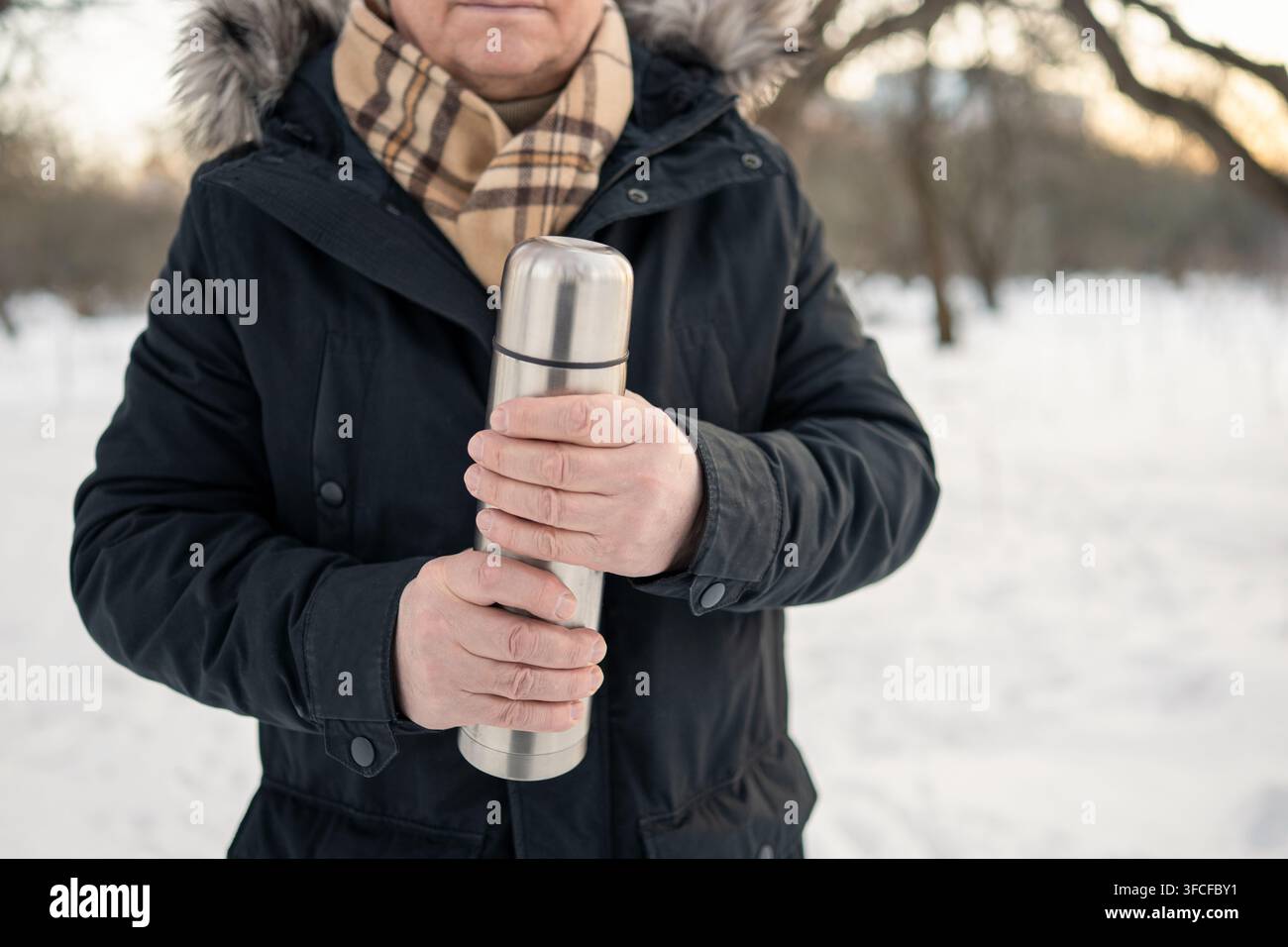 Anziano che tiene un thermos in acciaio inossidabile con entrambe le mani durante una passeggiata invernale in un parco innevato. Bevanda calda, freddo, invecchiamento attivo e stile di vita all'aperto Foto Stock