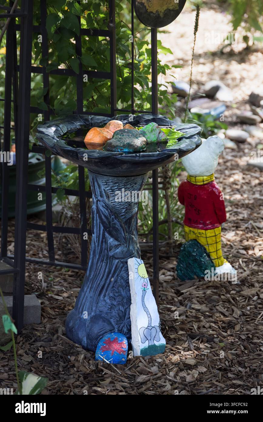 Una scena da giardino stravagante con una fontana per uccelli decorativa adornata con pietre dipinte colorate e un gnomo da giardino giocoso. L'ambientazione, situata a Cram Foto Stock