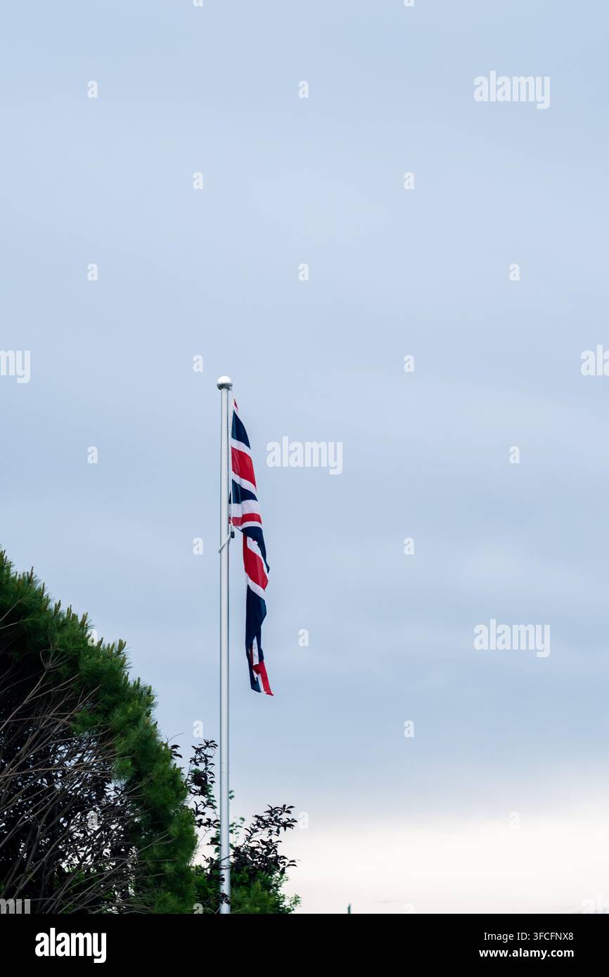 La bandiera britannica dell'Union Jack appesa a un alto asta su un cielo azzurro chiaro con rami di pino verdi in primo piano. 30 maggio 2025. Foto Stock
