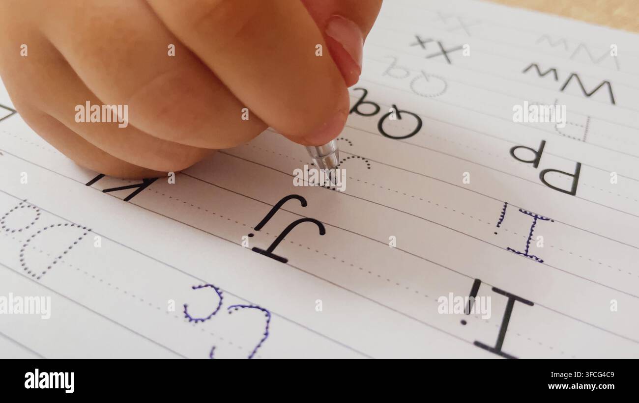 Vista macro in primo piano di una mano di bambino che tiene una penna tracciando le lettere J e I dell'alfabeto inglese puntinate in un quaderno di pratica della scrittura, foto stock orizzontale. Foto Stock