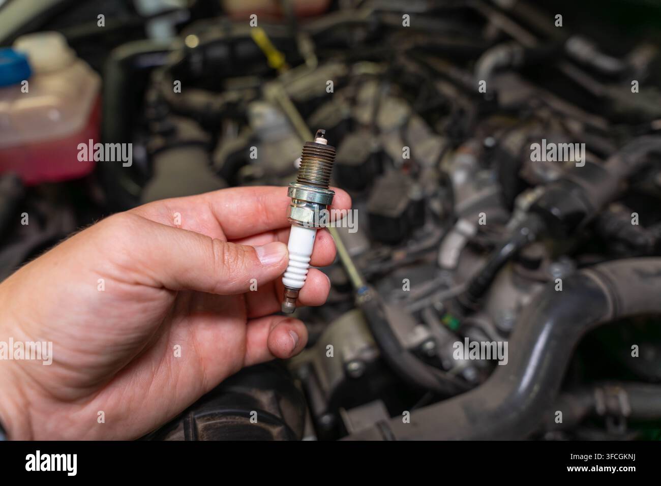 Candela di accensione sporca tenuta vicino al vano motore per la manutenzione del veicolo, una mano presenta la parte di accensione rimossa davanti a componenti meccanici, rendendo semplice Foto Stock
