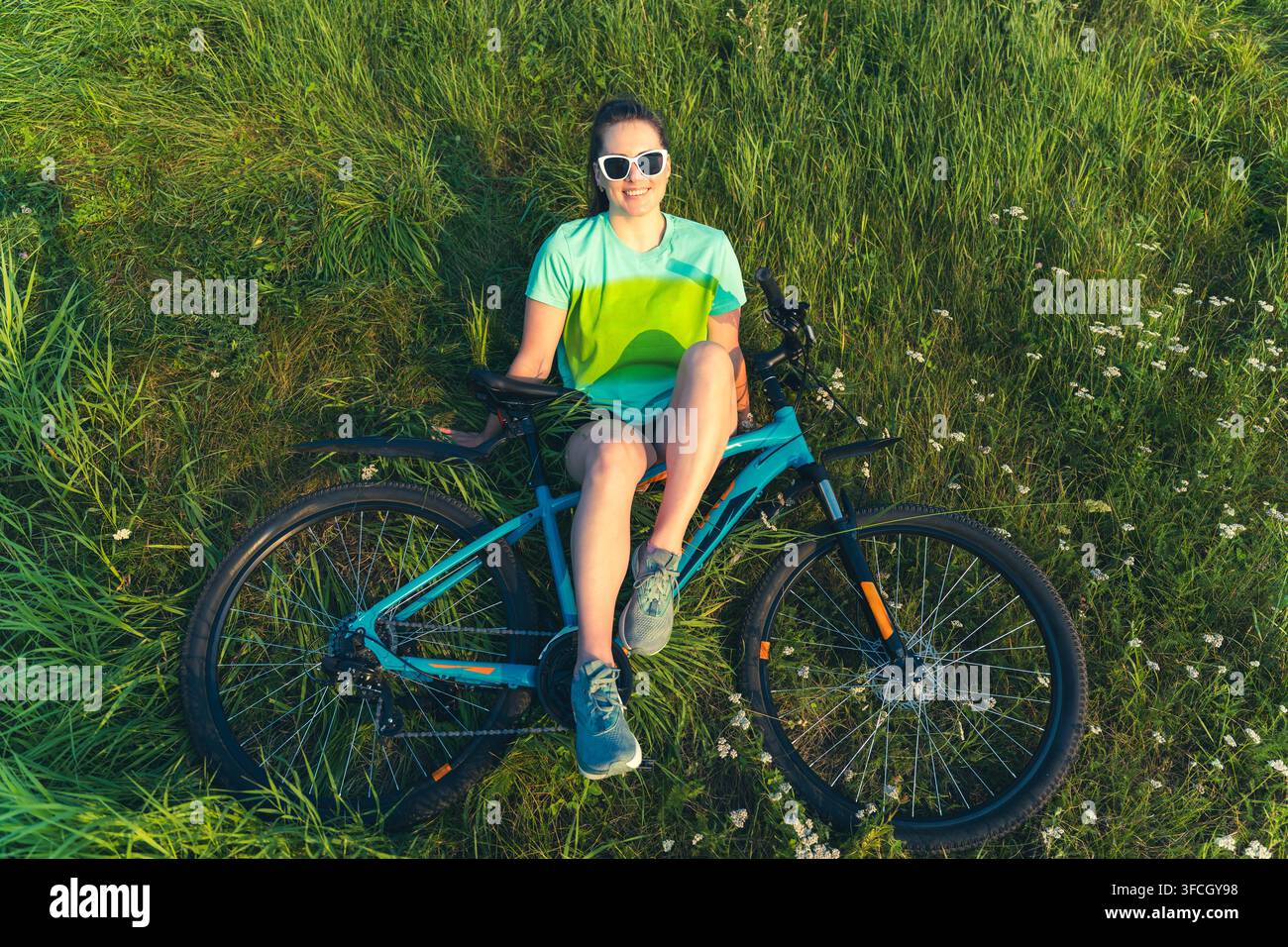 Ciclista sorridente seduta su una mountain bike in un prato verdeggiante, indossa occhiali da sole bianchi e una maglia sportiva. La sua posa rilassata Foto Stock