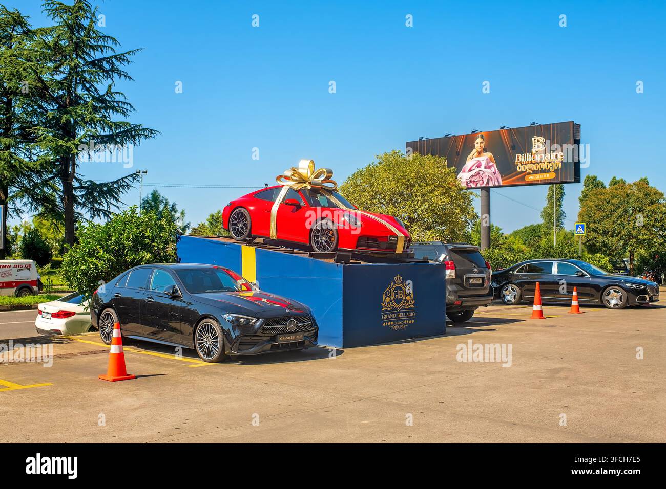 Batumi, Georgia - 22 agosto 2026: Auto sportiva rossa decorata con un grande fiocco dorato da regalo installata su un podio vicino a un casinò in una giornata soleggiata. Foto Stock