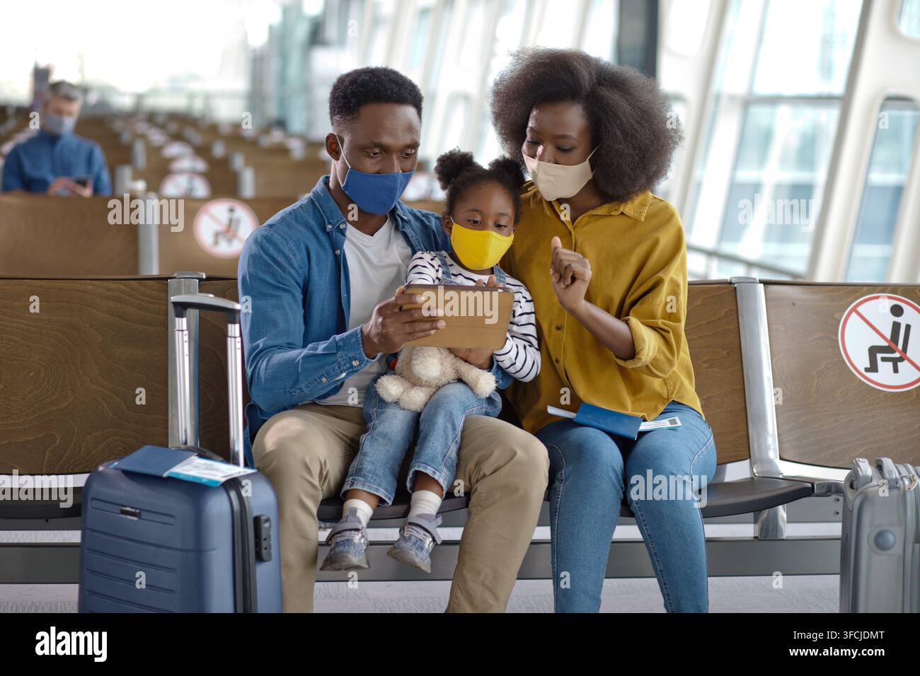 Un padre, una madre e una figlia sono seduti insieme in un terminal aeroportuale, tutti indossano coperture protettive per il viso e guardano un dispositivo digitale Foto Stock