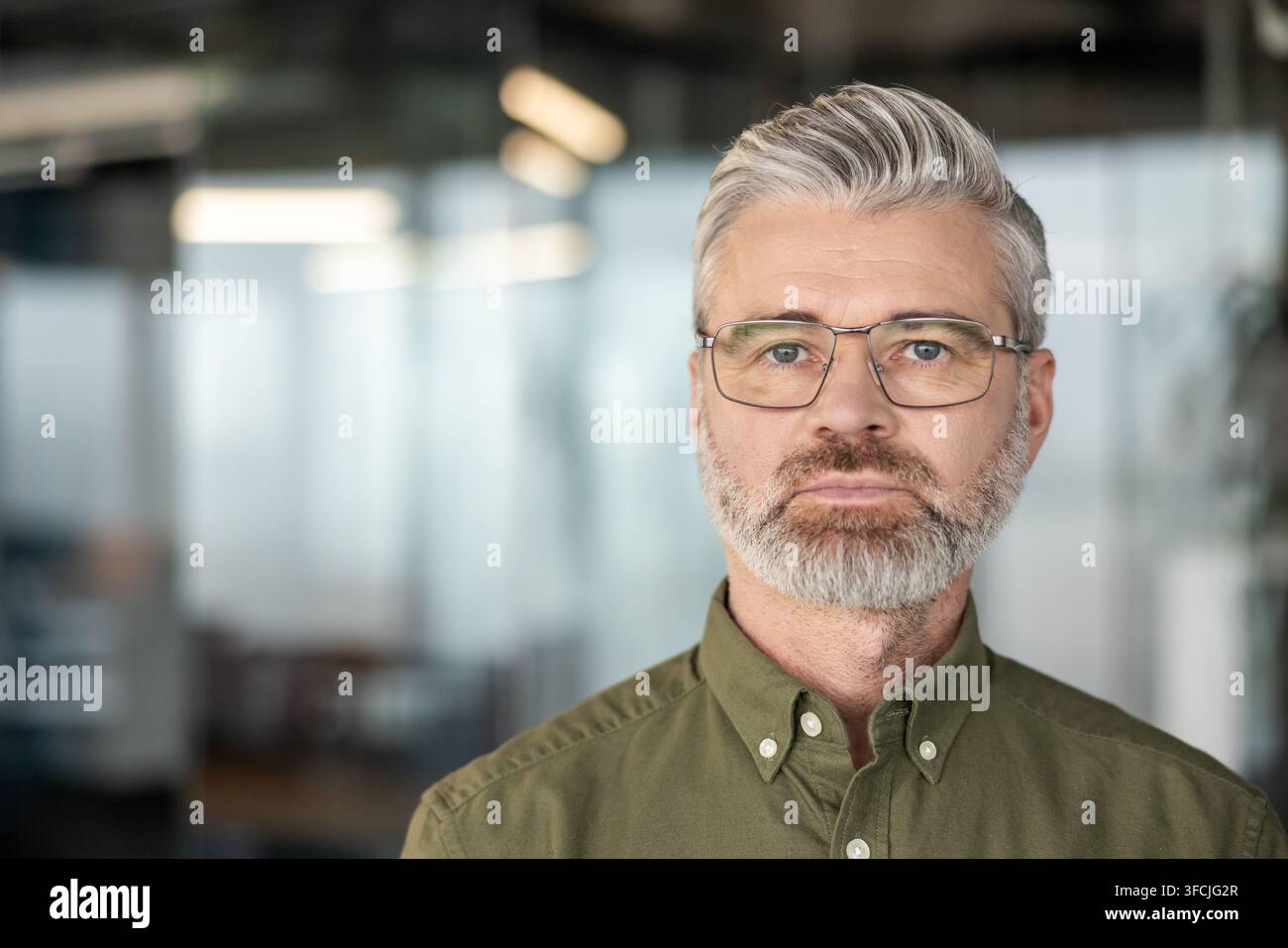 Uomo d'affari maturo serio con capelli grigi e barba, che indossa occhiali e una camicia verde, in piedi dalla vita in su in un ufficio moderno, sguardo diretto che trasmette sicurezza e leadership riflessiva Foto Stock