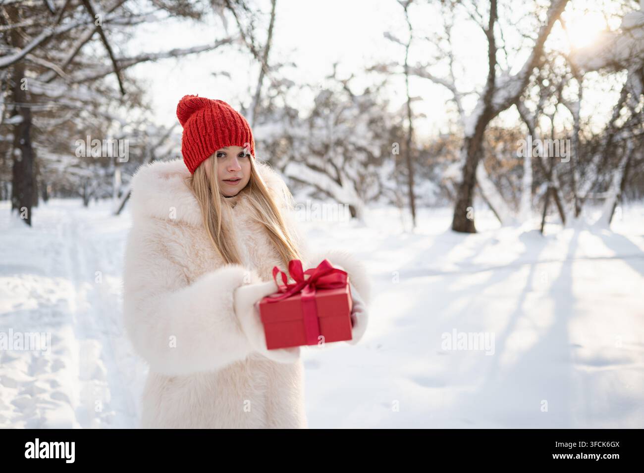 Giovane donna che tiene un regalo di Natale rosso verso la fotocamera in una foresta innevata, concetto di regalo festivo con luce invernale brillante e spazio per il testo. Foto Stock